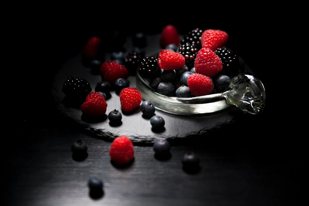 Mixed fresh berries, including raspberries and blueberries, in a glass bowl on a dark surface with scattered berries around.