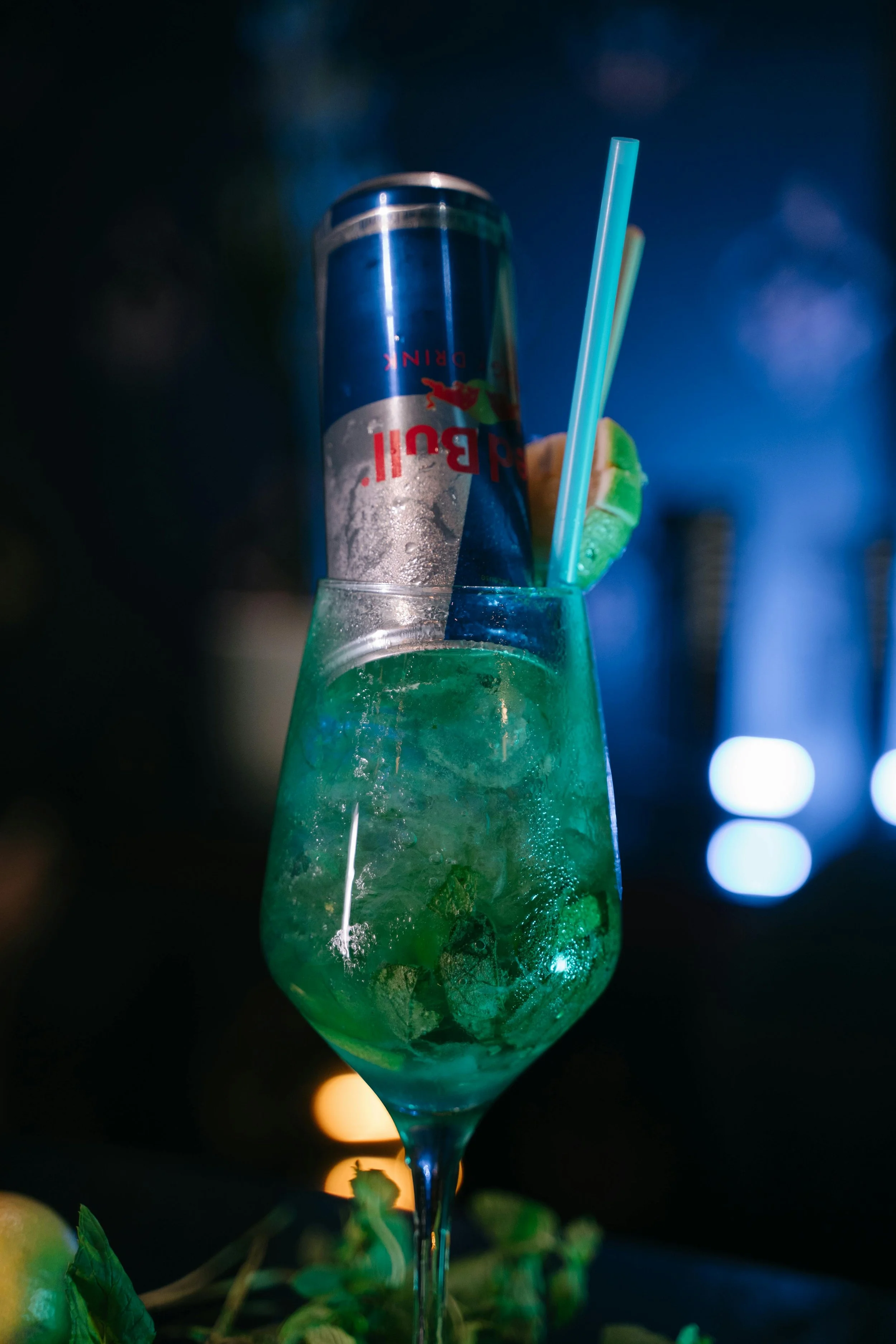 A cocktail glass with mint leaves and ice, with a can of Red Bull and two straws inside, set against a dark background with blue lighting.