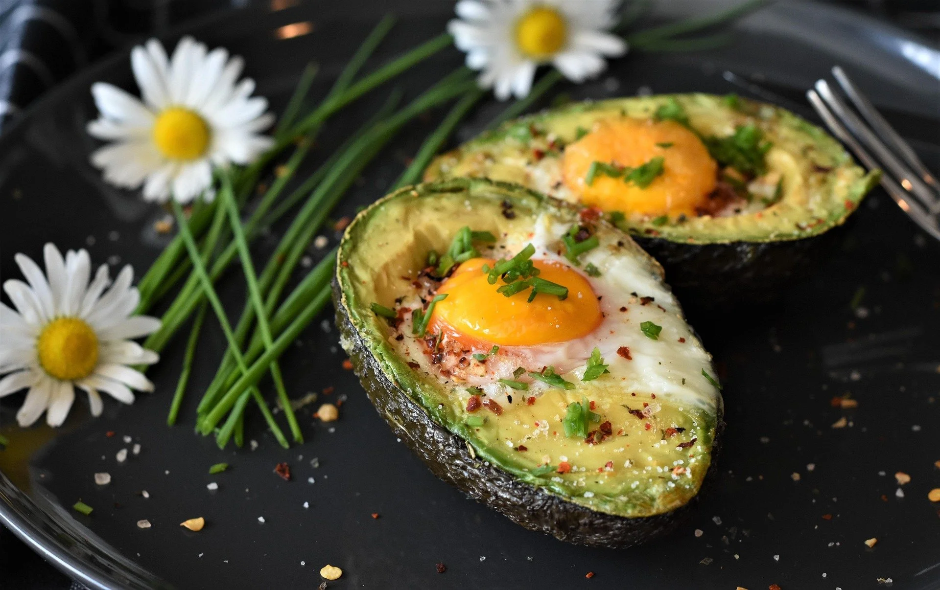 Baked avocado halves with eggs, garnished with chopped chives, served on a black plate with white daisy flowers and green herbs.