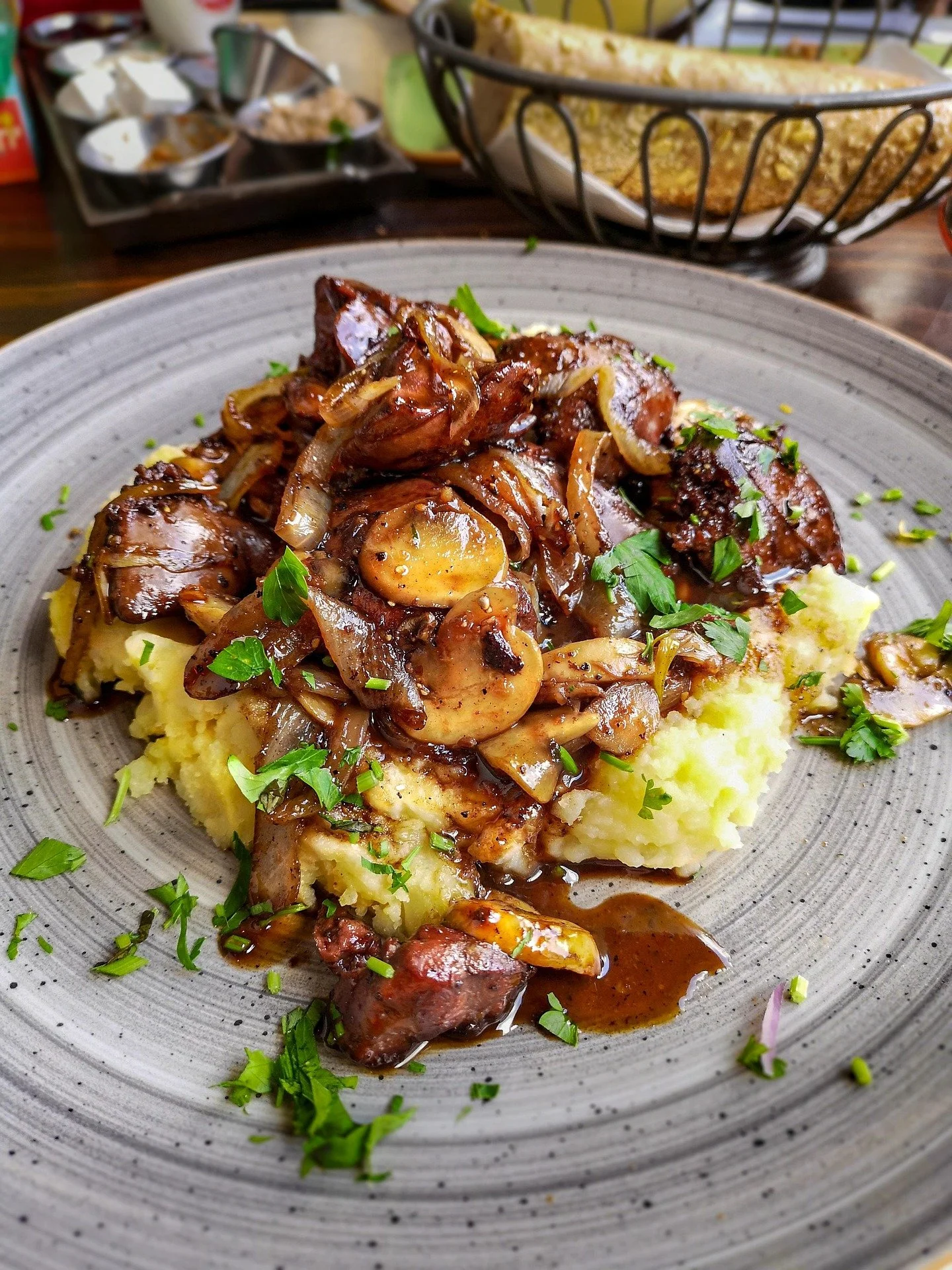 Plate of mashed potatoes topped with mushroom gravy, sautéed mushrooms, onions, and herbs, served with a basket of bread in the background.