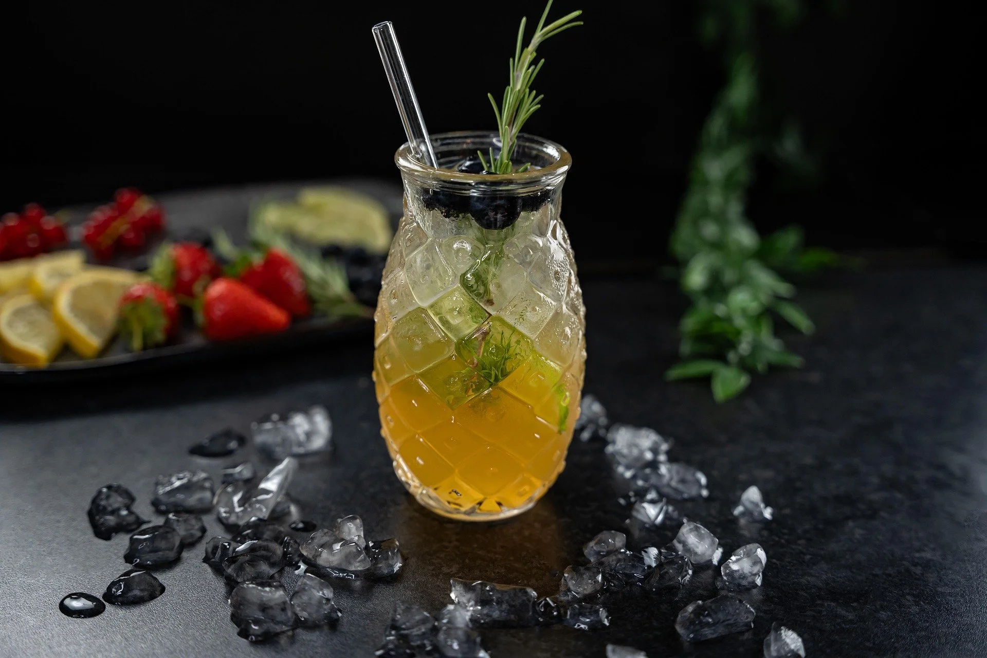 A glass pineapple-shaped container filled with a yellow cocktail, ice, lime slices, and garnished with rosemary and black olives, placed on a black surface with scattered ice around it.