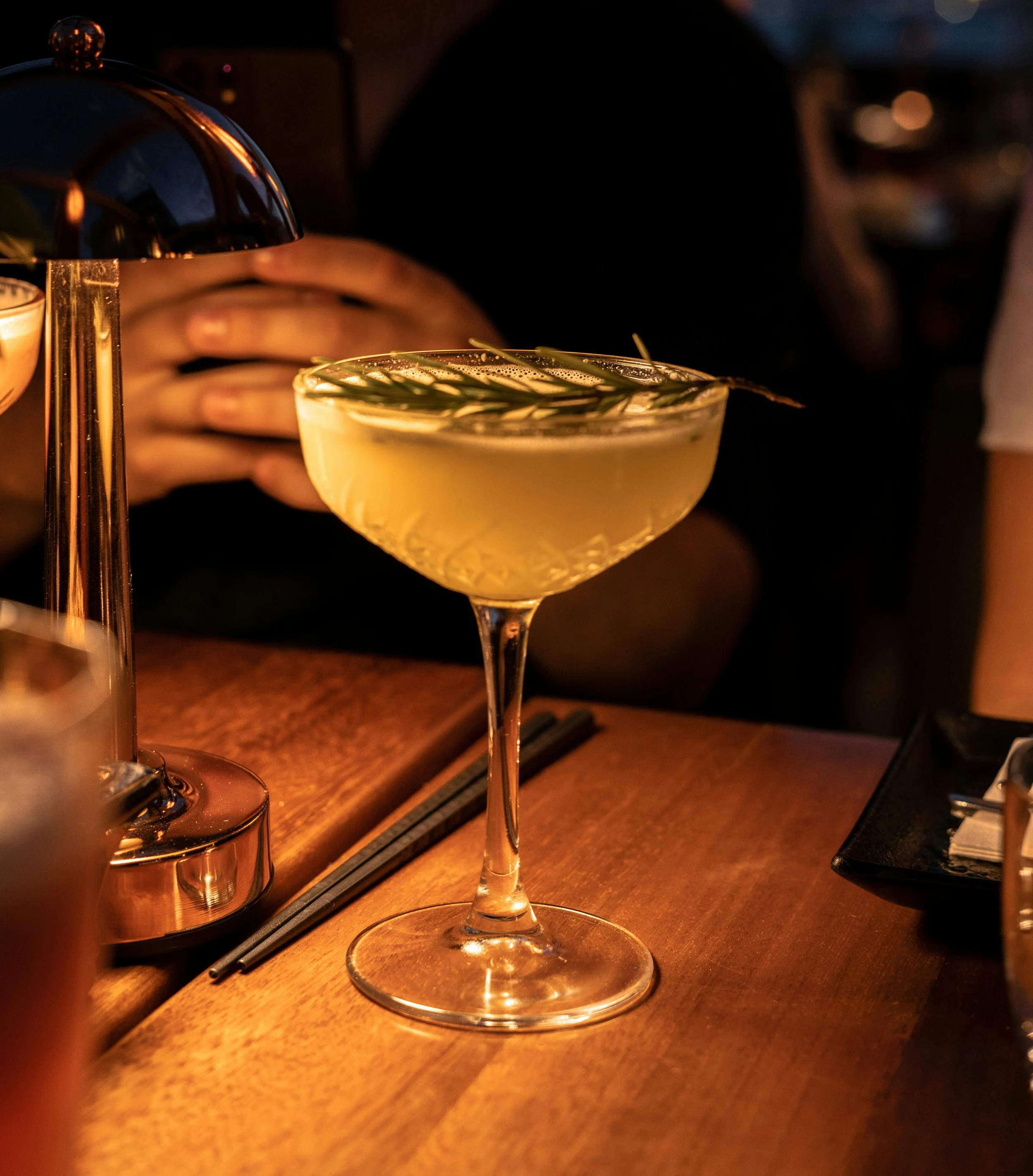 A cocktail drink in a cocktail glass garnished with herbs on a wooden table, with blurred background of people and ambient lighting.