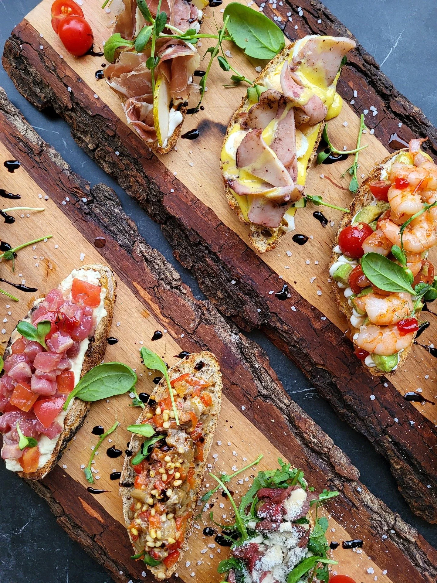 Assorted gourmet crostinis with various toppings such as prosciutto, shrimp, diced tomatoes, and cheese, garnished with microgreens, served on a wooden board with salt, pepper, and balsamic drizzle.