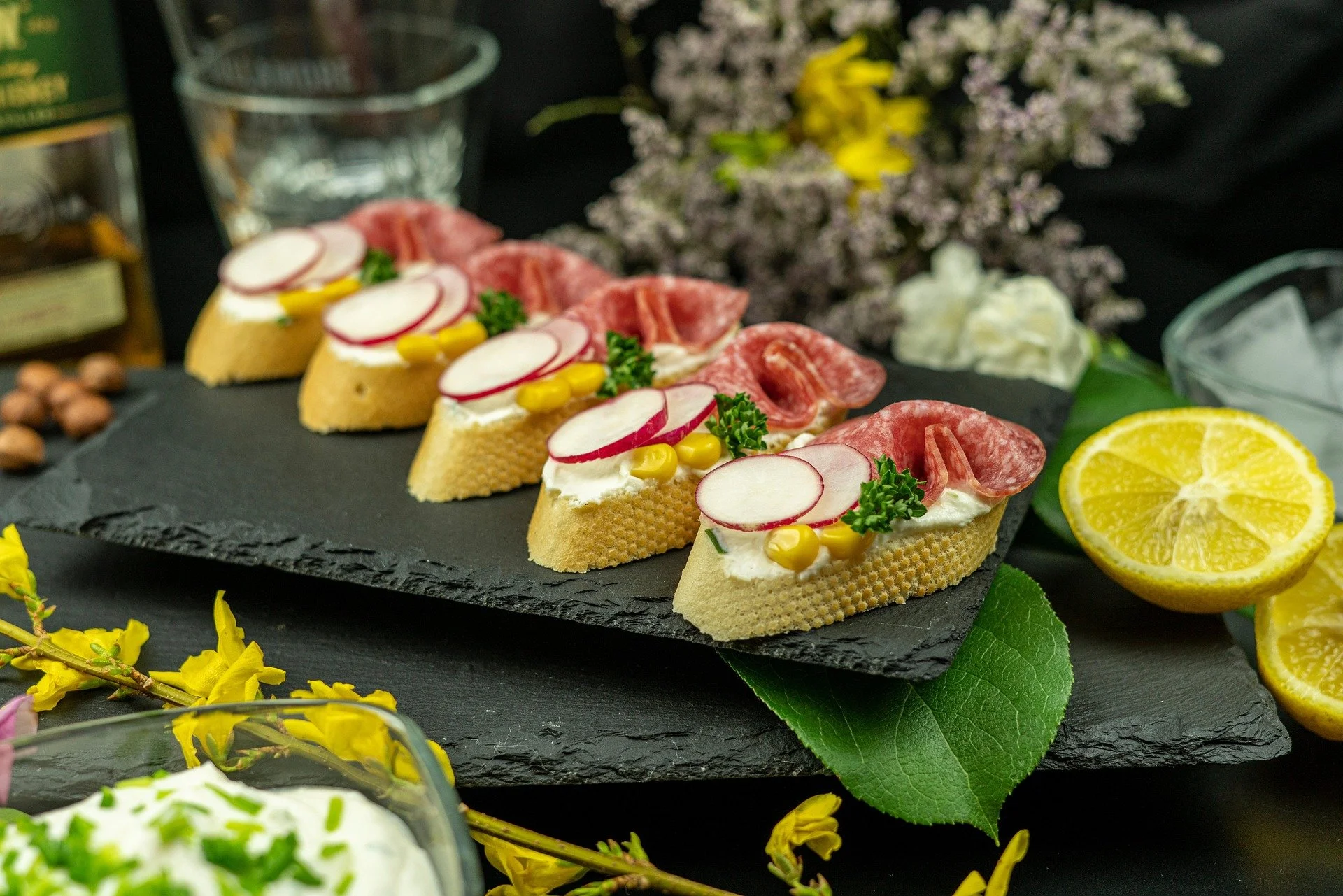 Canapés topped with radish slices, yellow corn, parsley, salami, and cheese on a black slate platter, with lemon half and garnishes in the background.