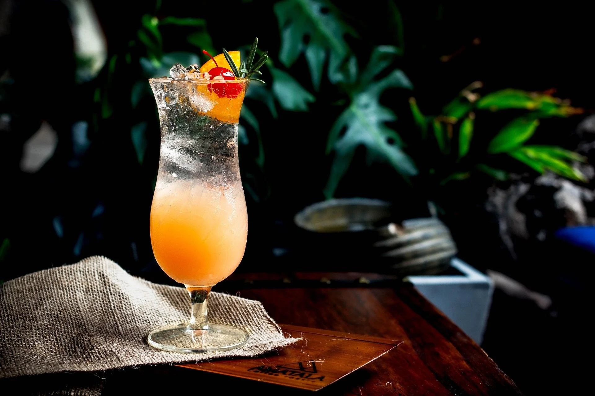 A tropical cocktail in a tall, curvy glass with orange juice, ice, garnished with cherry, lemon, and herbs, placed on a cloth on a wooden table.