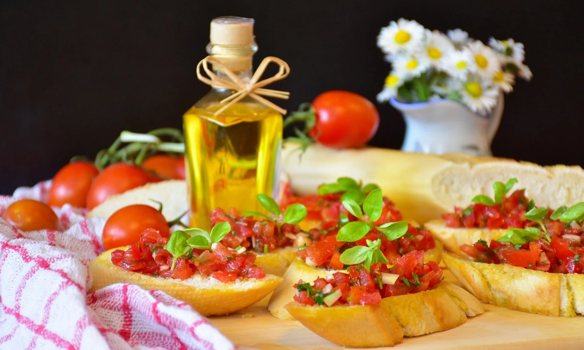 Bruschetta with tomato and basil on a wooden cutting board, cherry tomatoes, a bottle of olive oil, and daisies in a white pitcher.