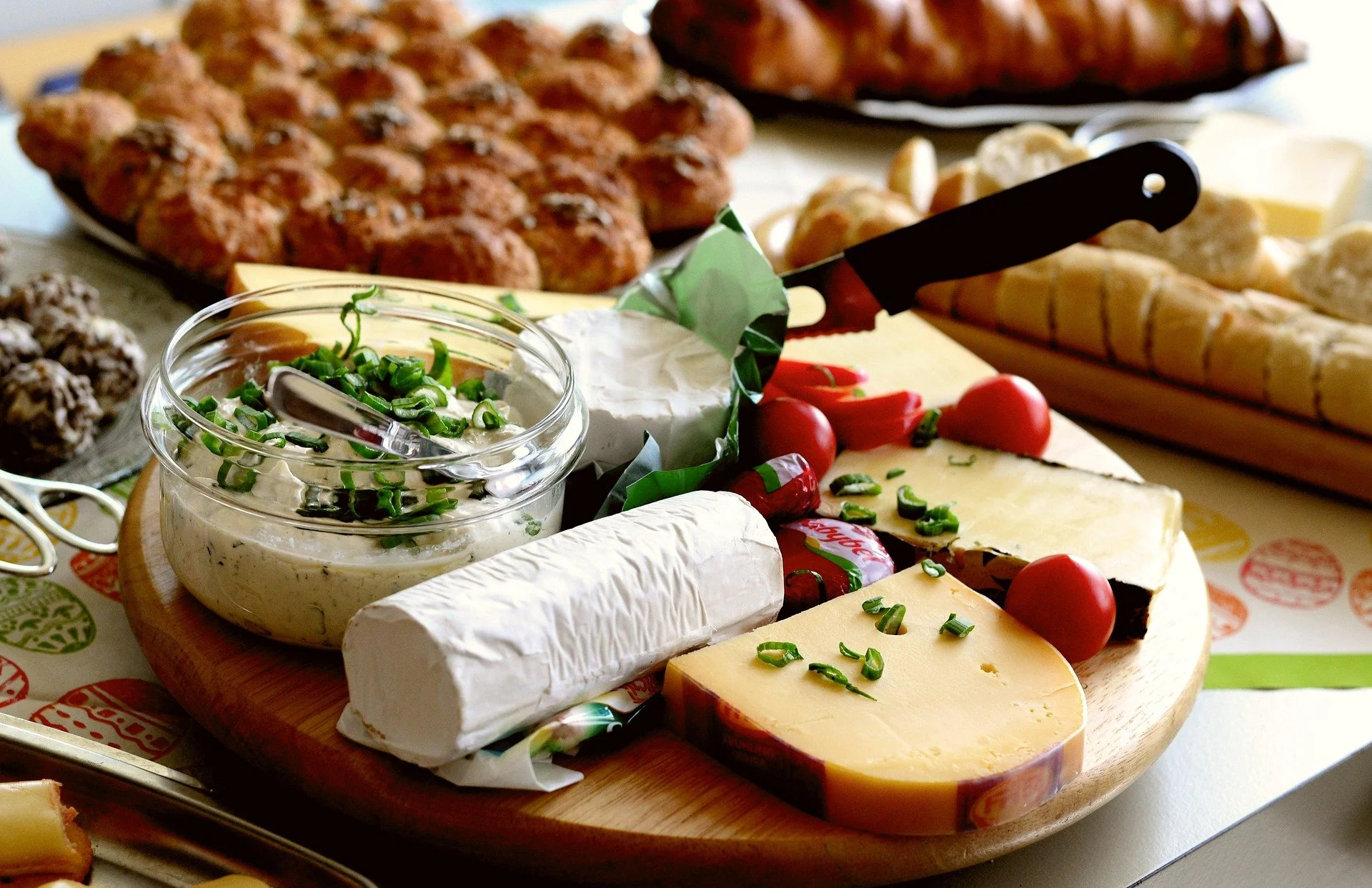 Cheese, cherry tomatoes, and a cheese spread on a cheese board with a background of baked goods.