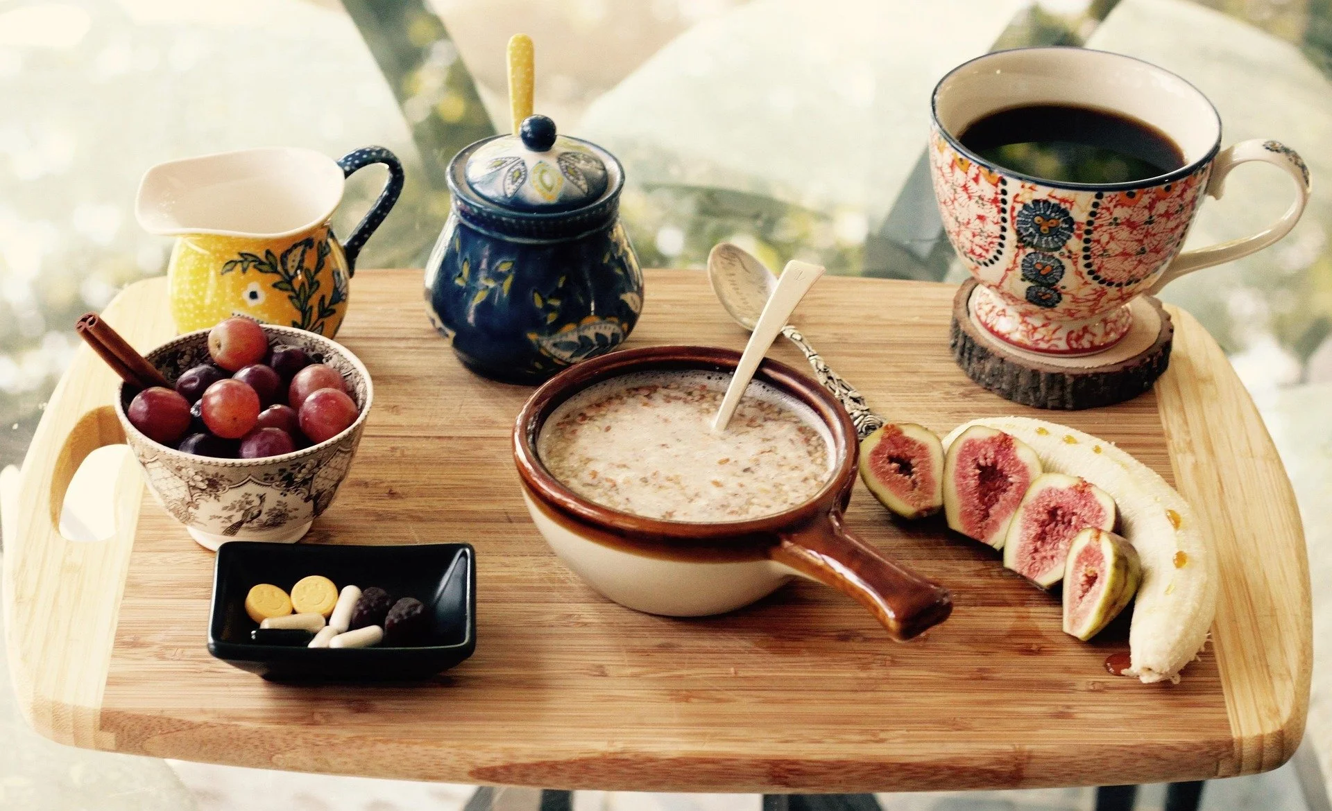A wooden tray holding a cup of coffee, a bowl of cereal, a small bowl of grapes, a small pitcher, a sugar bowl, a spoon, some sliced figs and a peeled banana, and a small dish of pills or vitamins.