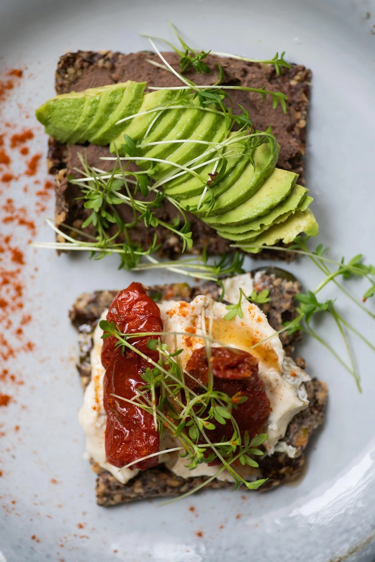 Close-up of two slices of avocado toast with microgreens, cherry tomatoes, poached egg, and seasoning on a white plate.