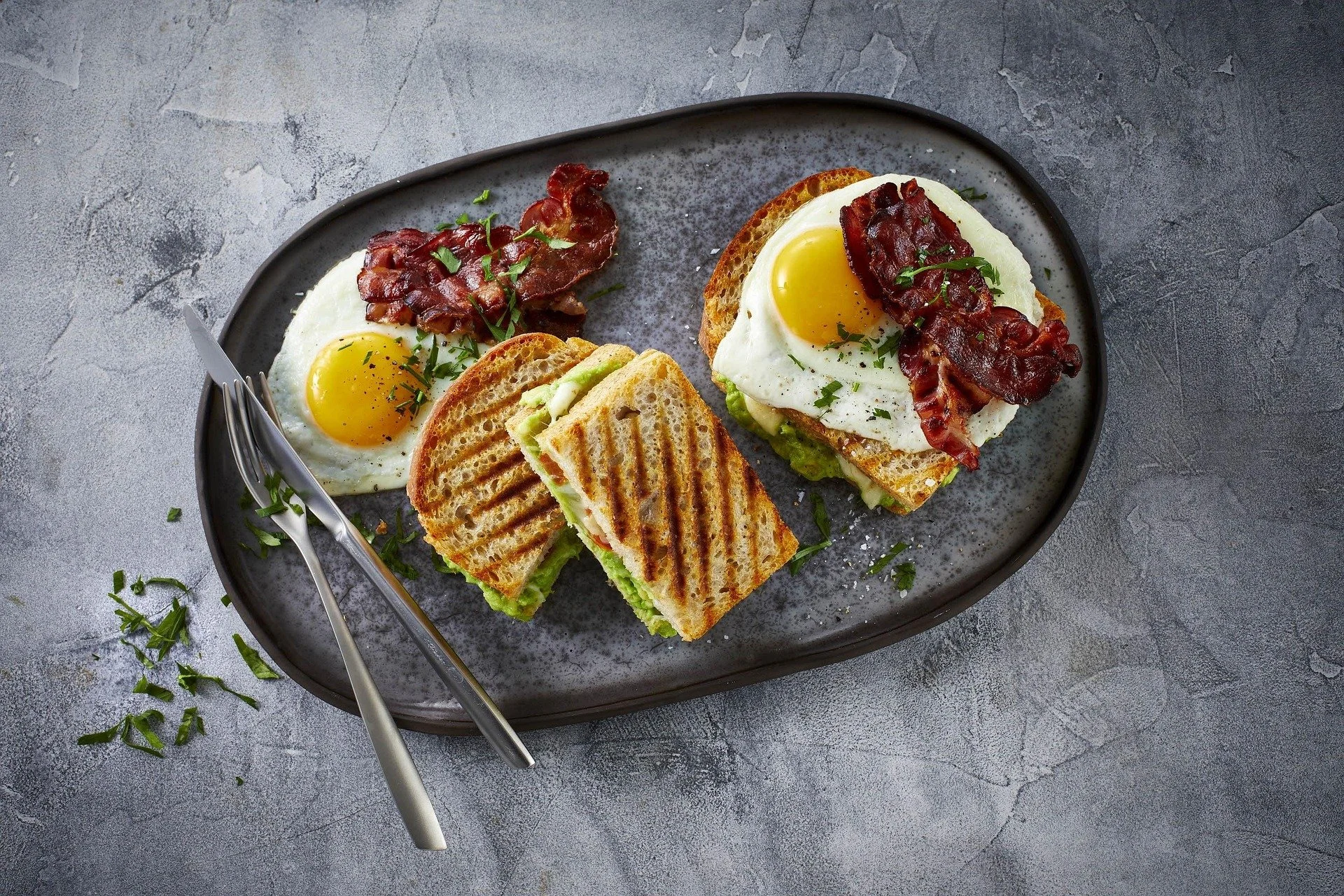 A black oval plate with two breakfast sandwiches and two sunny-side-up eggs with bacon. The sandwiches contain fried eggs on toasted bread with bacon and vegetables, garnished with chopped herbs. A fork and knife are placed on the plate, with some ch