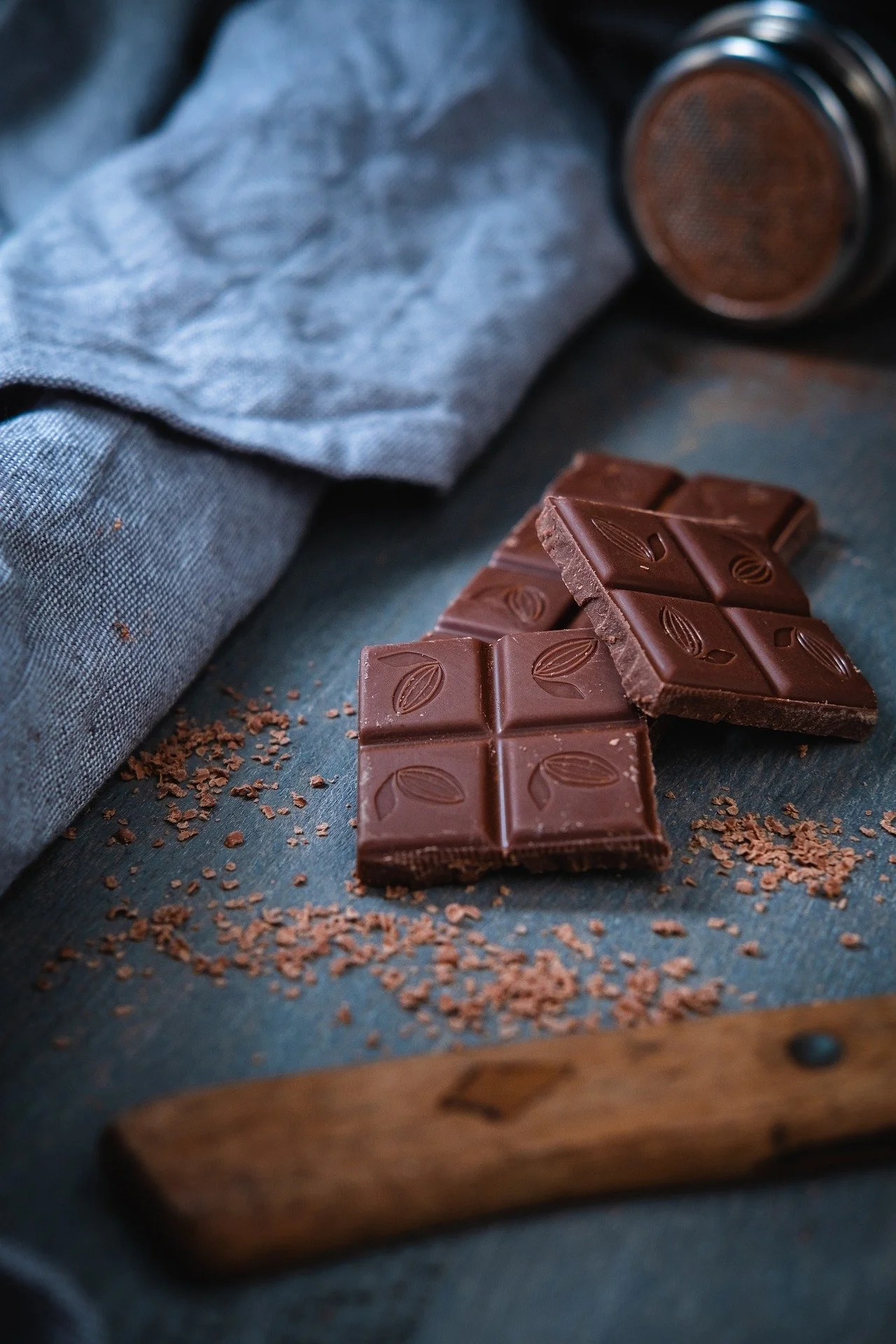 Pieces of milk chocolate with a leaf pattern on top, scattered cocoa powder, a blue cloth, and a blunt knife on a dark wooden surface.
