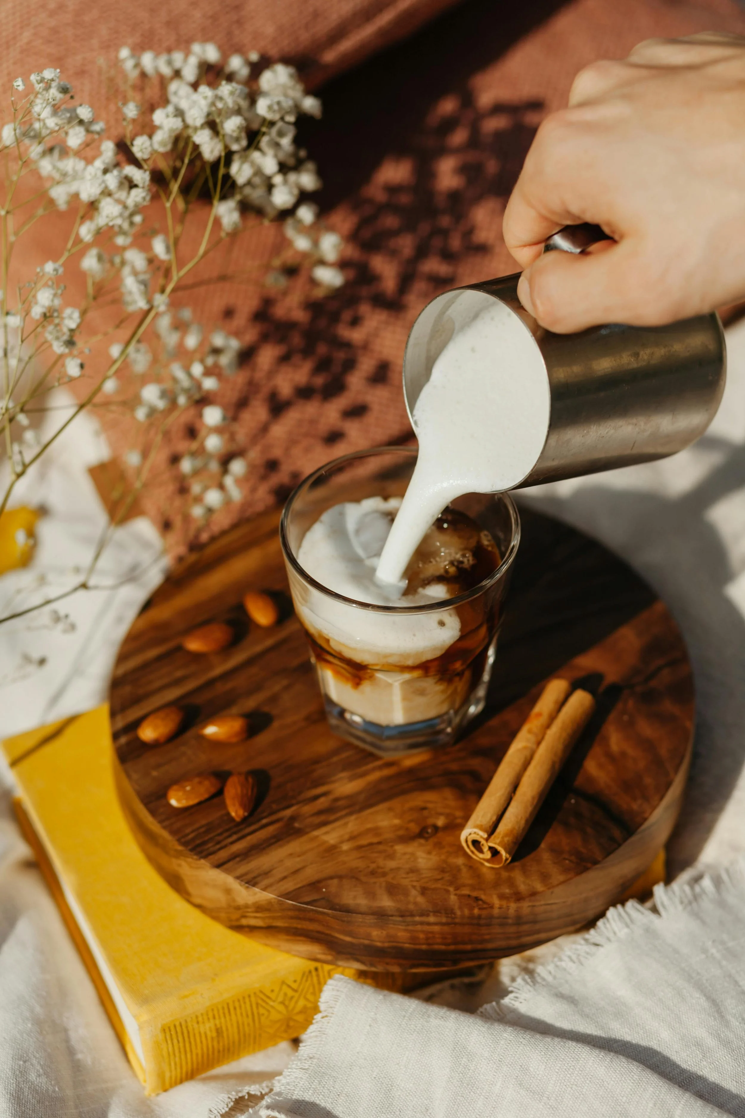 Pouring cream into iced coffee with cinnamon sticks and almonds on a wooden tray