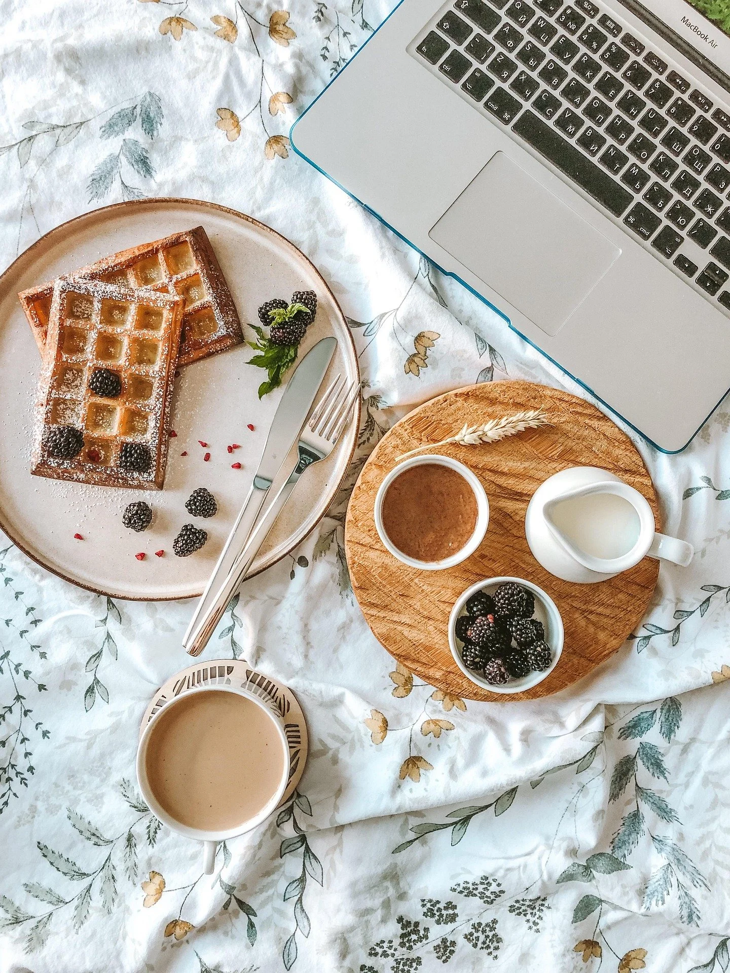 Breakfast setup with waffles topped with blackberries on a white plate, two cups of coffee, a small bowl of blackberries, a small jug of milk, a small container of chocolate spread, and a laptop on a floral bedsheet.