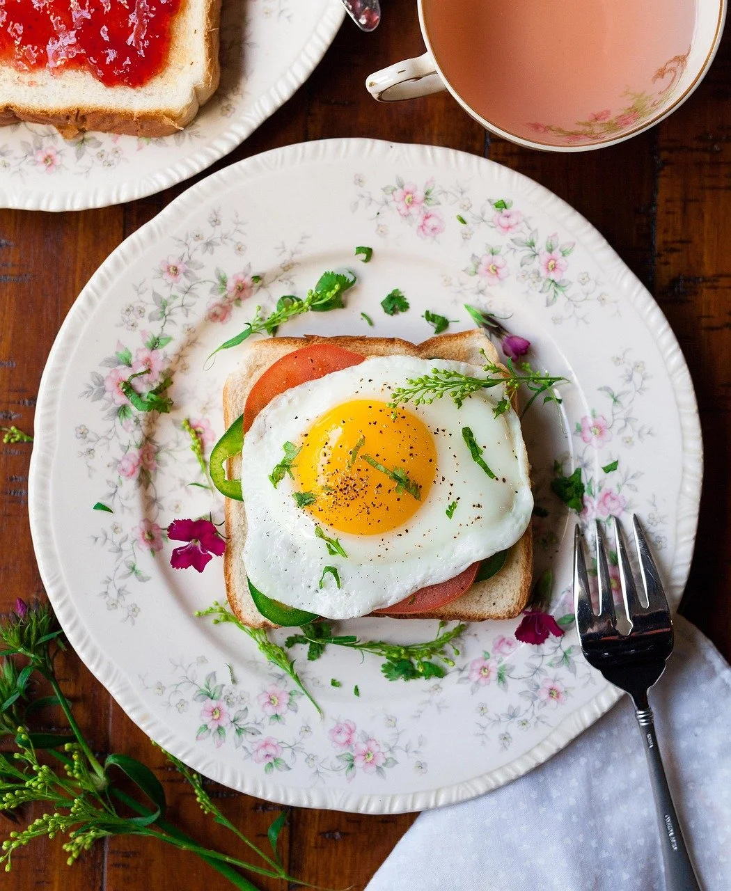 A plate with toast, topped with a fried egg, tomato, cucumber, and herbs, with a fork, a cup of tea, and a jar of jam on the side.
