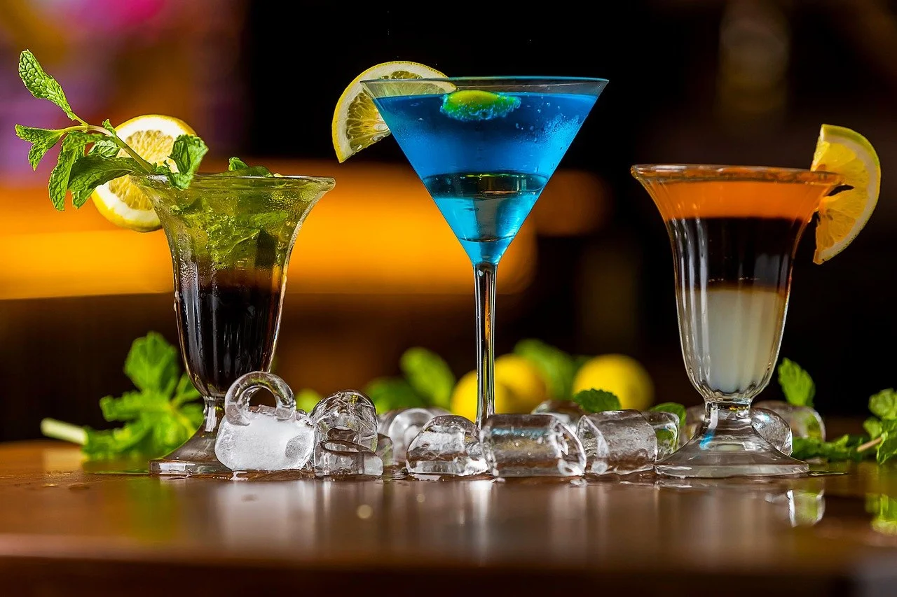 Three colorful cocktails on a wooden surface with ice cubes, lemon slices, and mint leaves, illuminated against a dark background.