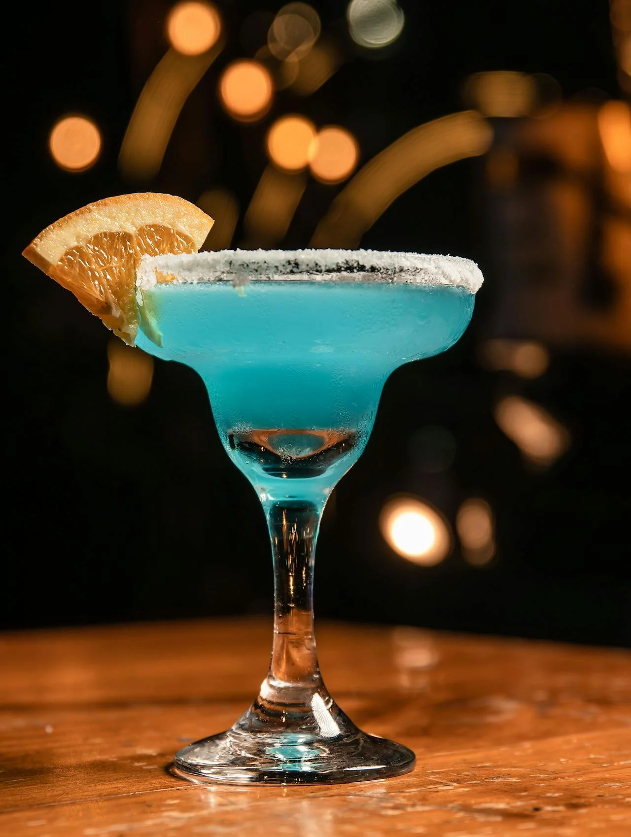 A blue cocktail in a Margarita glass garnished with an orange wedge and lemon wedge, with a salted rim, on a wooden surface with blurred warm lights in the background.