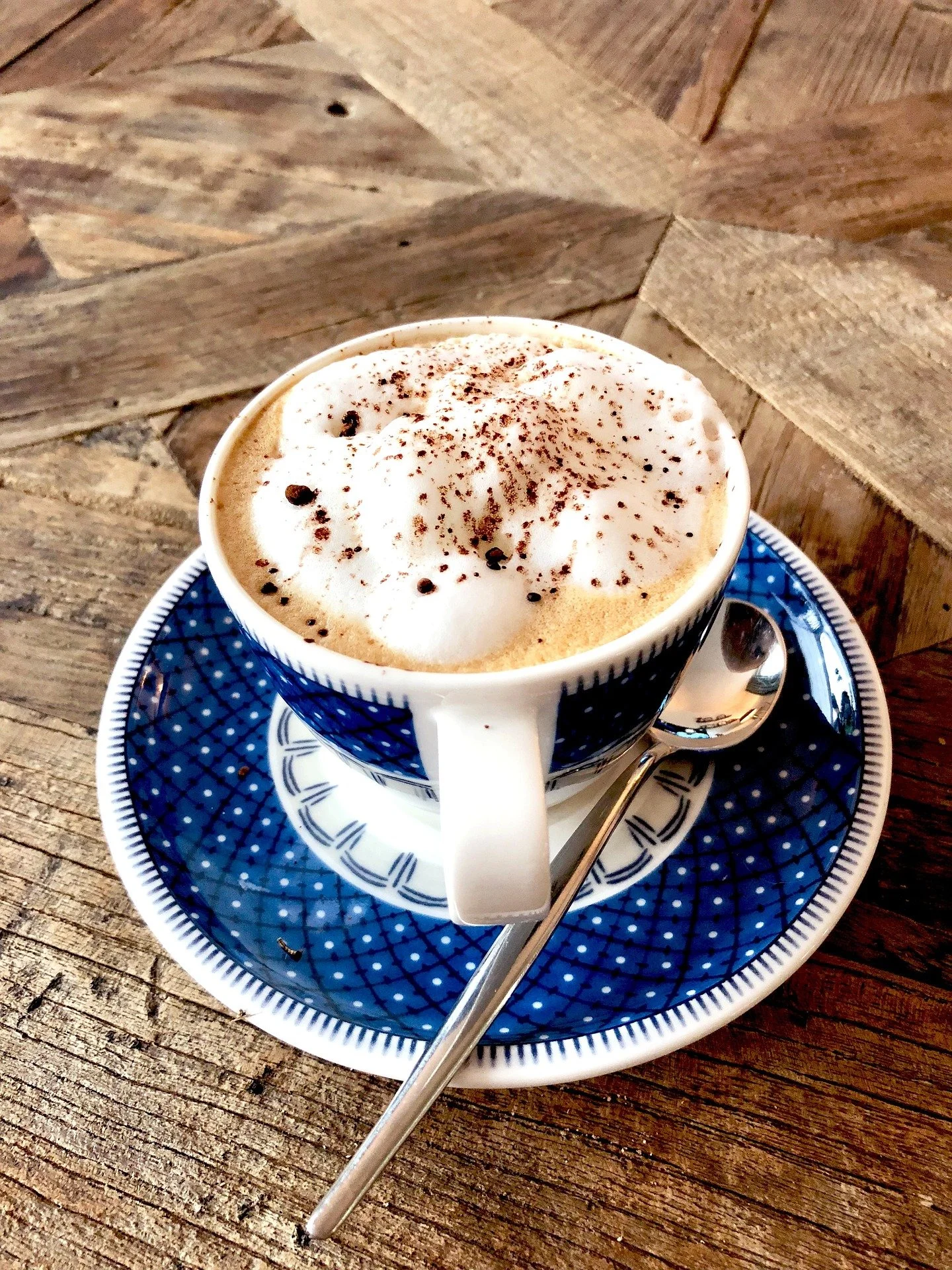 A cup of coffee topped with whipped cream and cocoa powder, served on a blue patterned saucer with a silver spoon on a rustic wooden table.