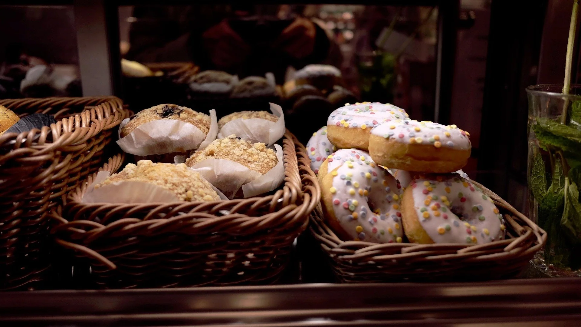 Wicker baskets containing muffins and donuts with colorful sprinkles, displayed behind a glass in a bakery or café setting.
