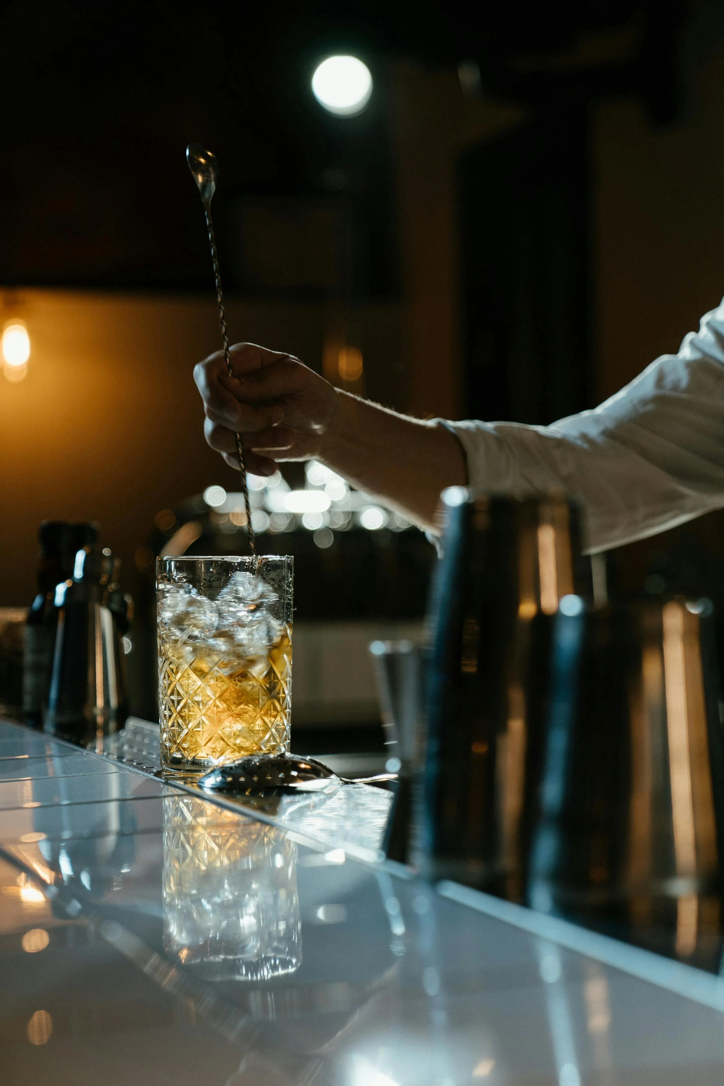 A bartender stirring a cocktail in a glass with ice, in a dimly lit bar setting.