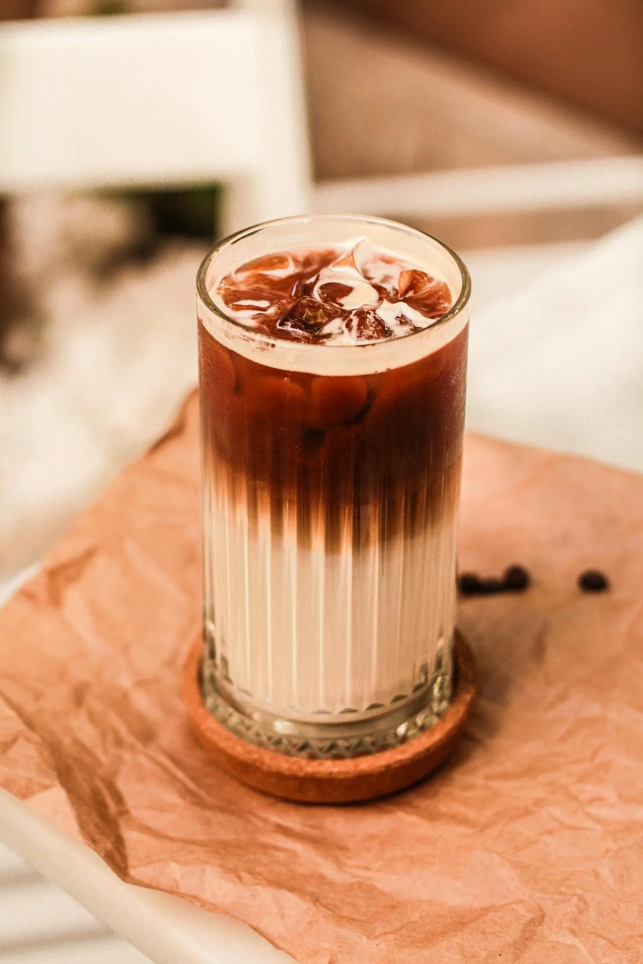A tall glass of iced coffee with layers of cream and coffee, served on a small round wooden coaster on a pinkish paper napkin.