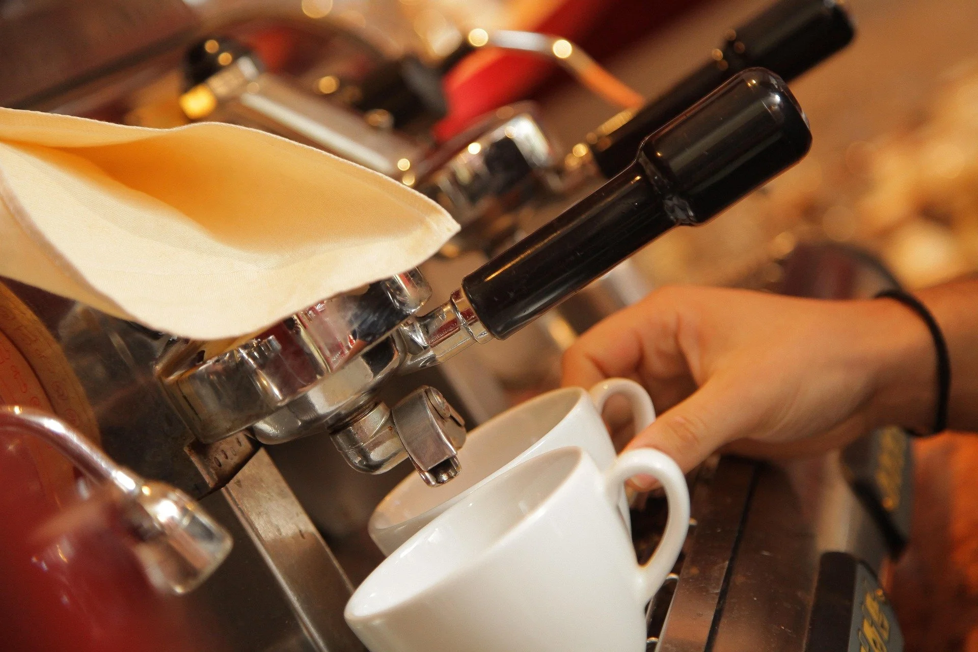 Close-up of a barista removing espresso shot cups from a coffee machine with a hand, and a white cup being filled.