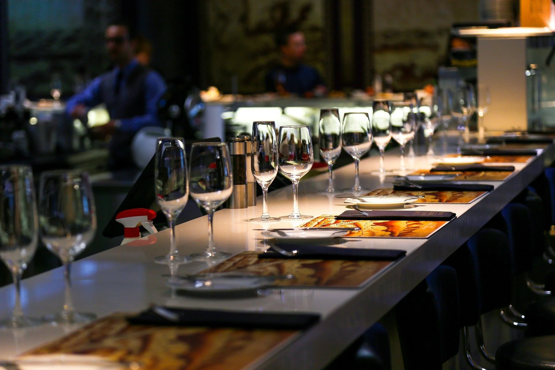 A row of empty wine glasses and place mats with cutlery on a restaurant bar counter with blurred background of staff and kitchen area.