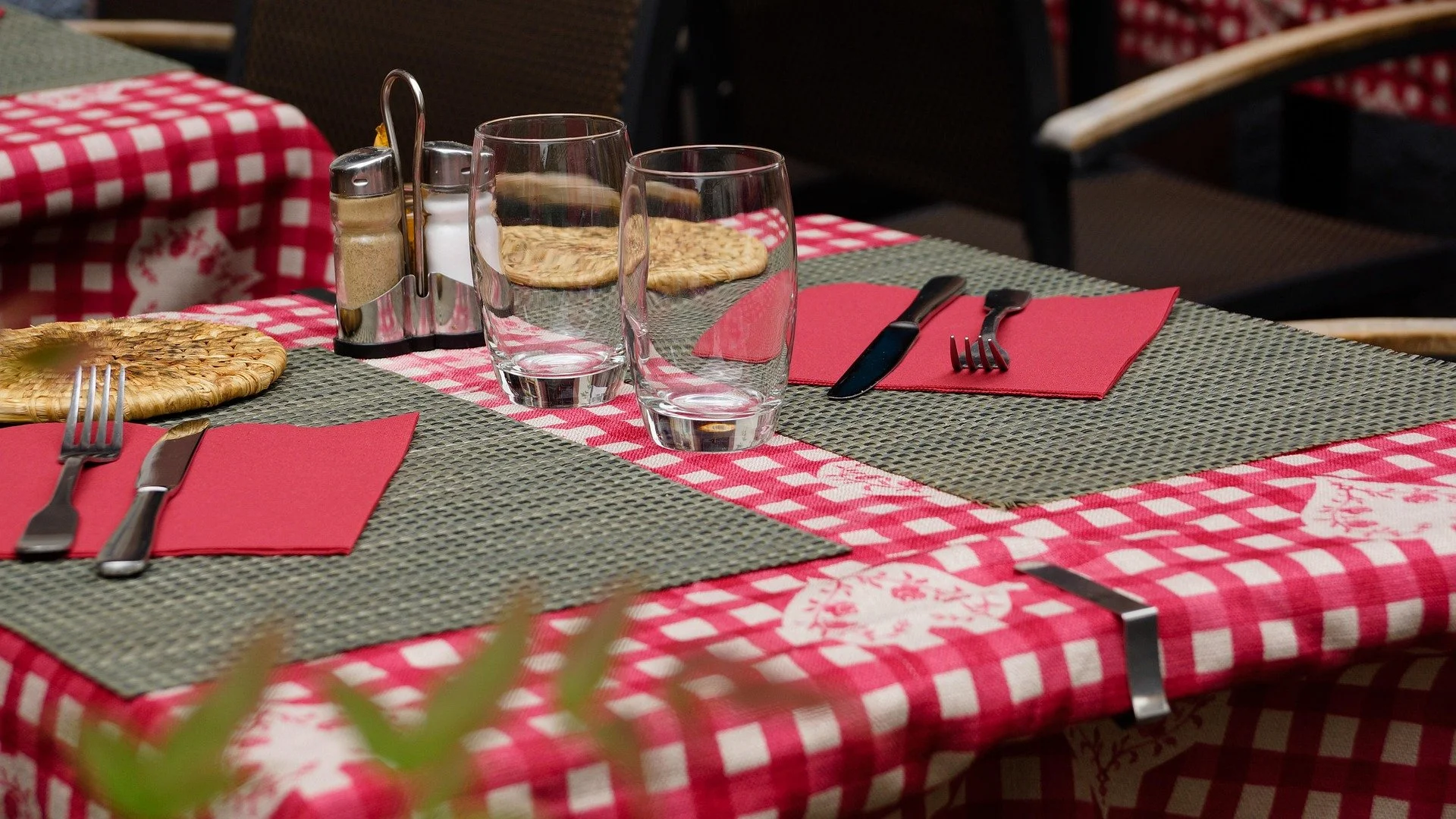 A dining table set with red and white checkered tablecloth, woven placemats, plates, glasses, utensils on red napkins, and a salt and pepper shaker, in an outdoor restaurant setting.