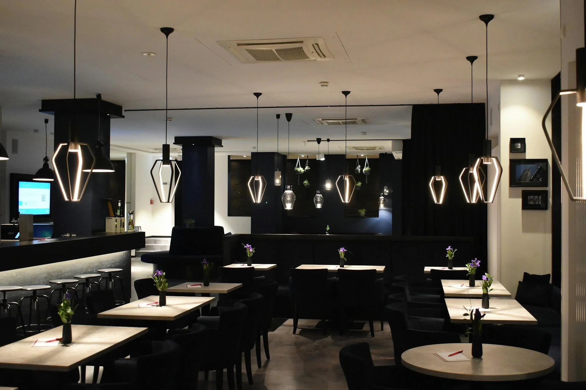 Interior of a modern restaurant or cafe with black and white decor, hanging pendant lights, tables with vases of purple flowers, and a bar area on the left.