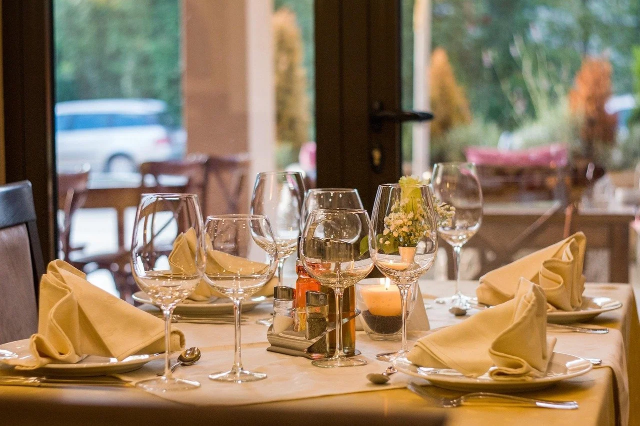 A table set for a meal with wine glasses, napkins, silverware, a candle, and a small flower arrangement near a window with a view outside.