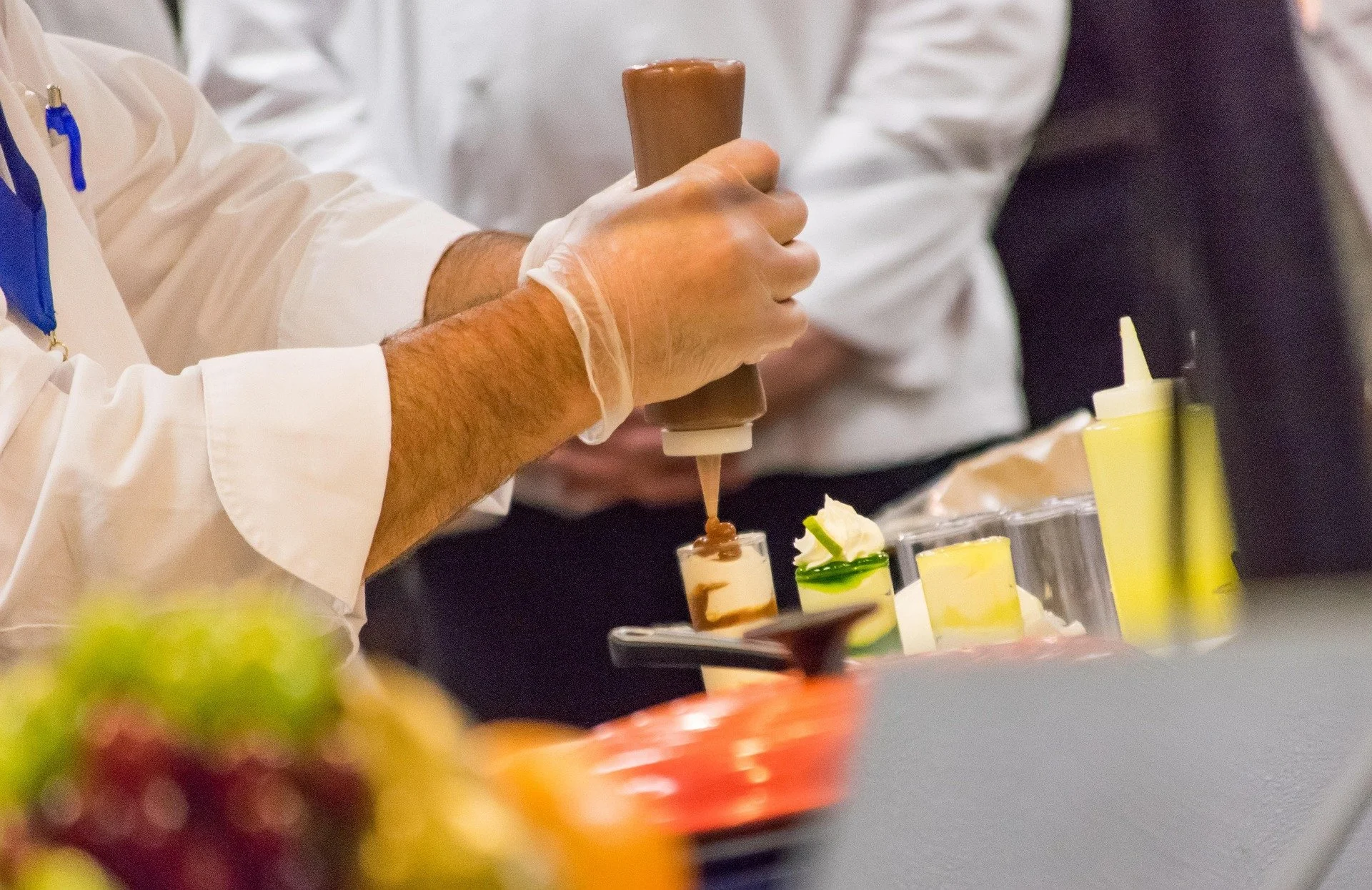 A chef in a white coat is decorating desserts with chocolate using a squeeze bottle. Various colorful desserts and toppings are on the table.