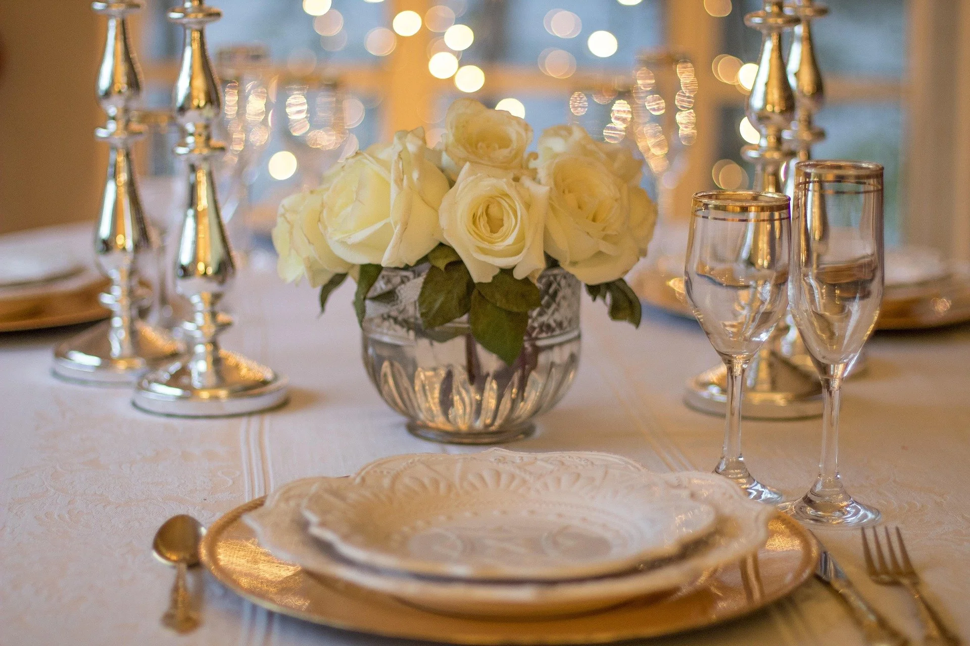Elegant dining table set with white roses in a silver vase, gold-rimmed glassware, and fine china while soft lighting creates a sophisticated ambiance.