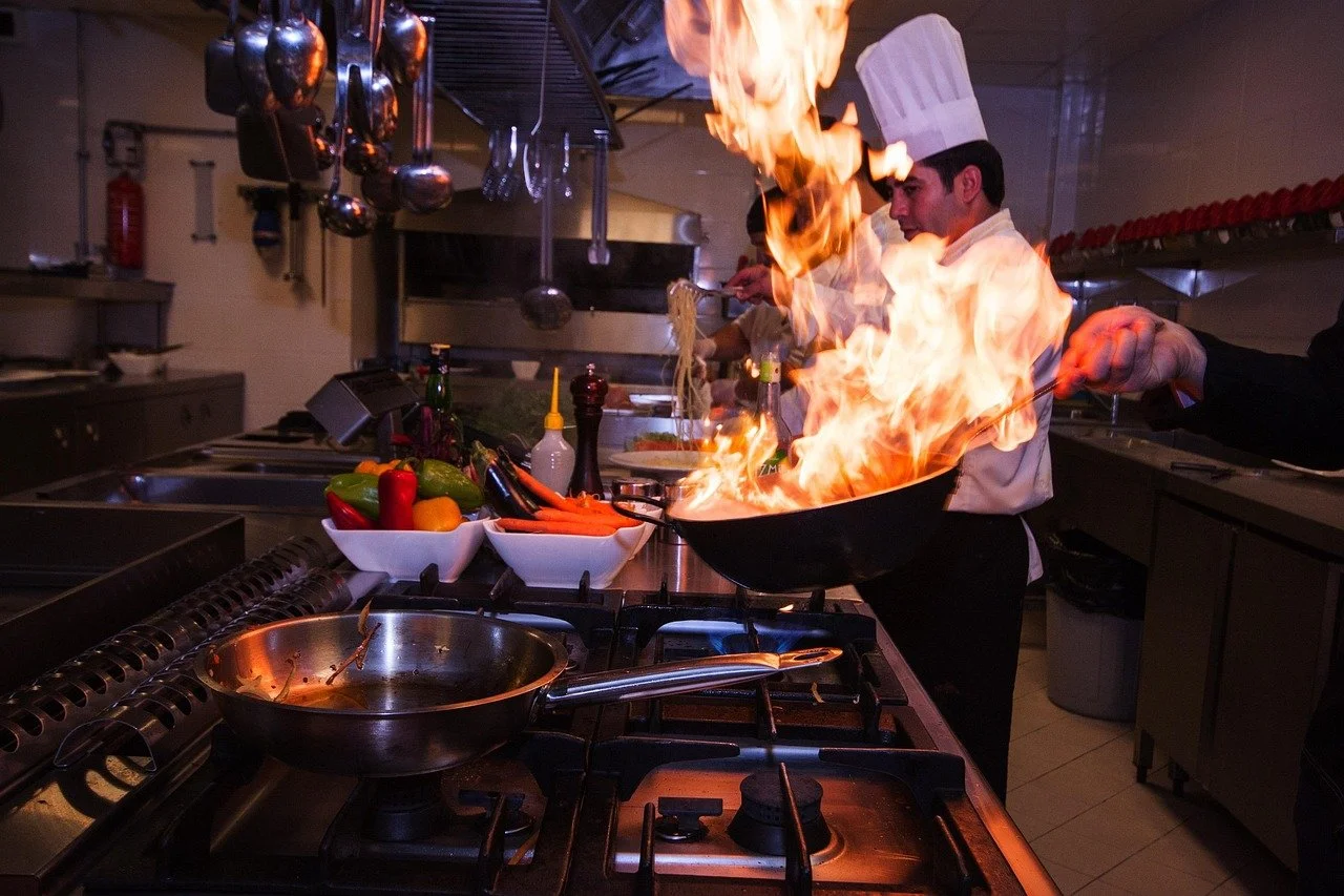 A chef wearing a white hat flaming a wok on a busy kitchen stove with various kitchen tools, vegetables, and condiments on the counter.