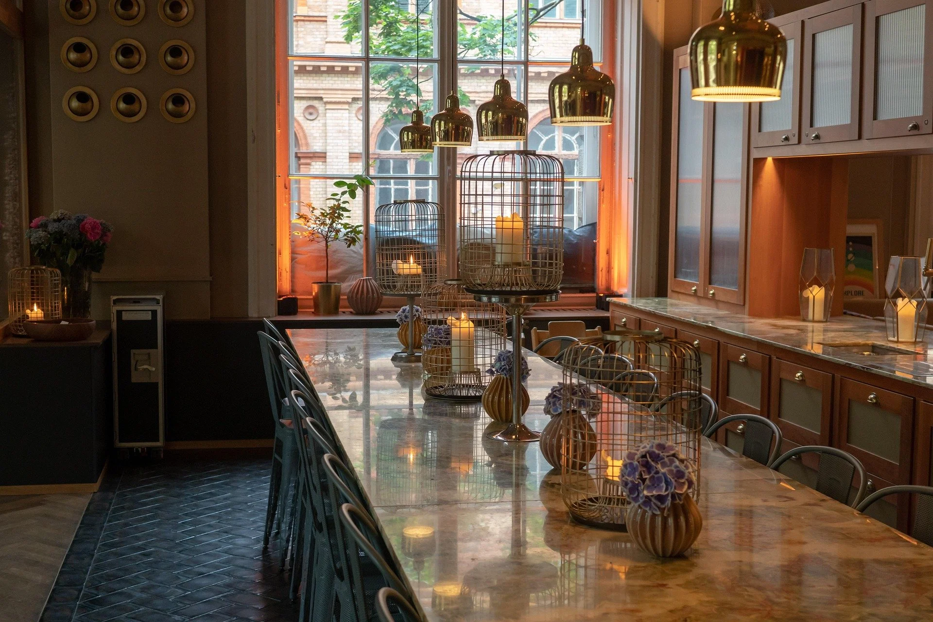 A long dining table with decorative candle cages and flower vases, set in a warmly lit room with large windows, hanging gold pendant lights, wooden cabinets, and a view of brick buildings outside.
