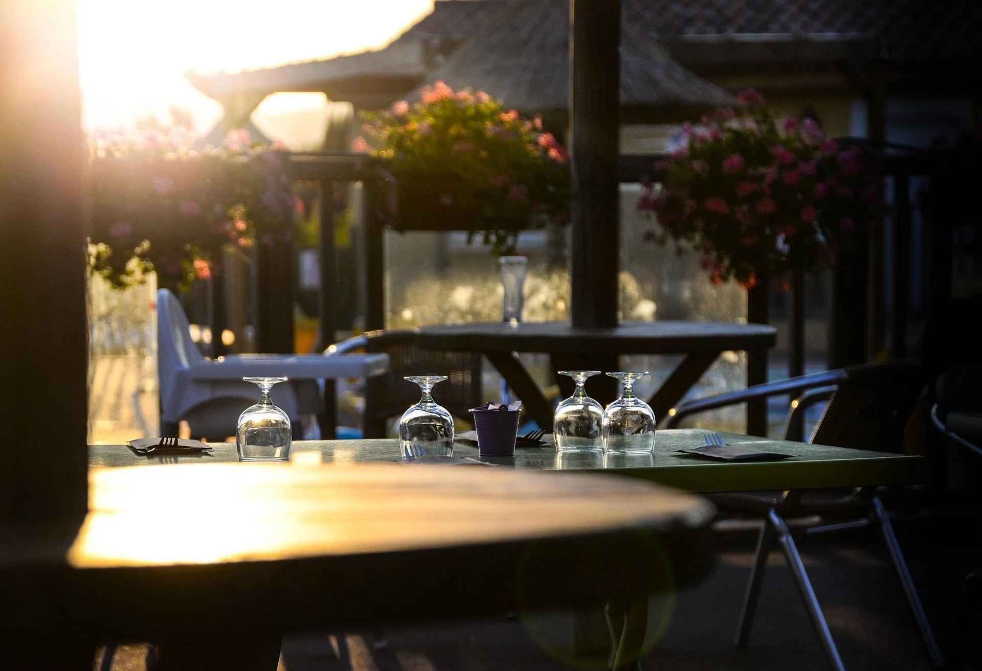 Empty outdoor restaurant table set with upside-down wine glasses, with sunlight and flowers in the background.