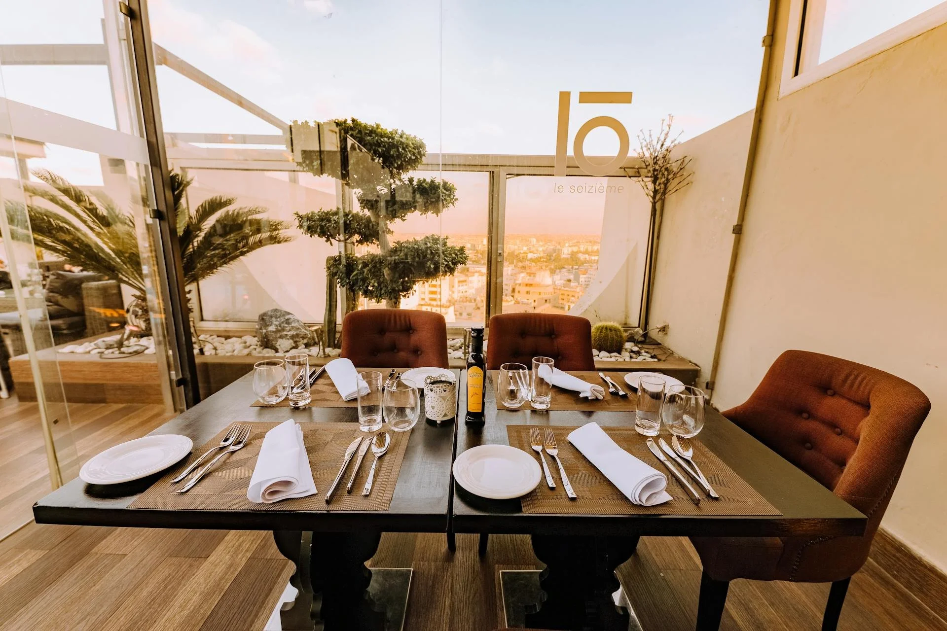 A restaurant table set for four people with white plates, silverware, white napkins, and glasses, on a dark wood table with brown placemats, in front of a glass wall with a view of a city skyline at sunset, decorative plants and rocks in the backgrou