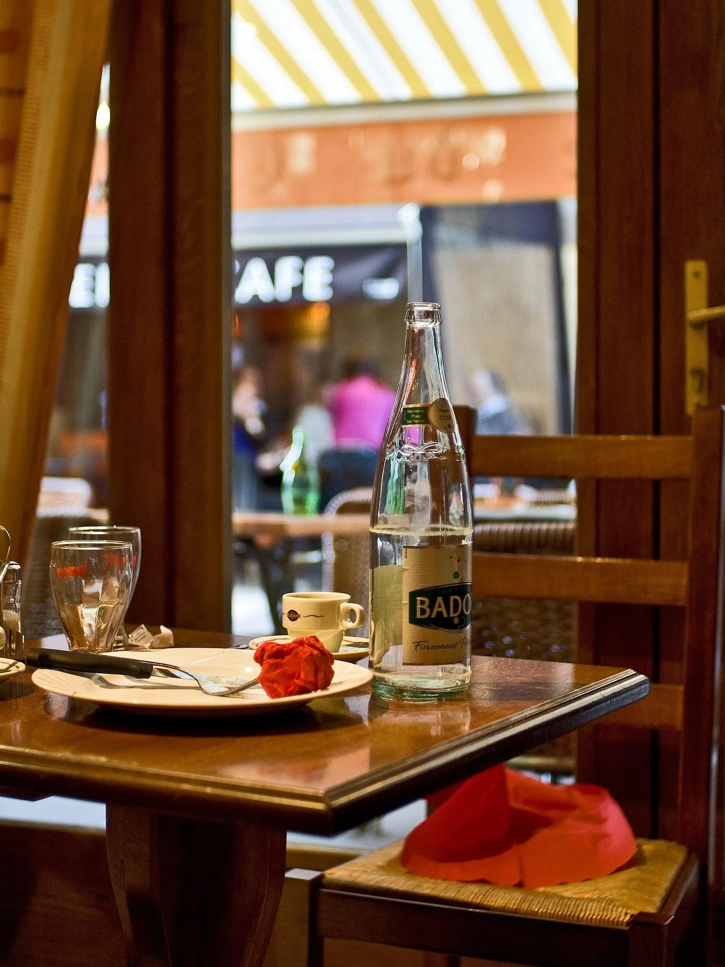 A table inside a restaurant with a glass bottle of water, a small cup, a glass of water, and a plate with utensils and a piece of bread. The table is next to a window showing a blurry outdoor cafe scene with people and a black sign that reads 'CAFE'.