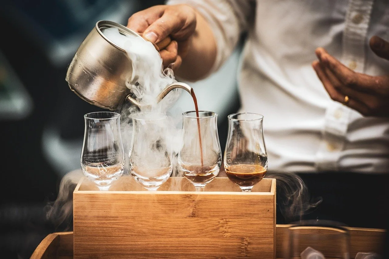 A person pouring hot coffee into glasses with steam rising, from a metal pot, on a wooden tray.