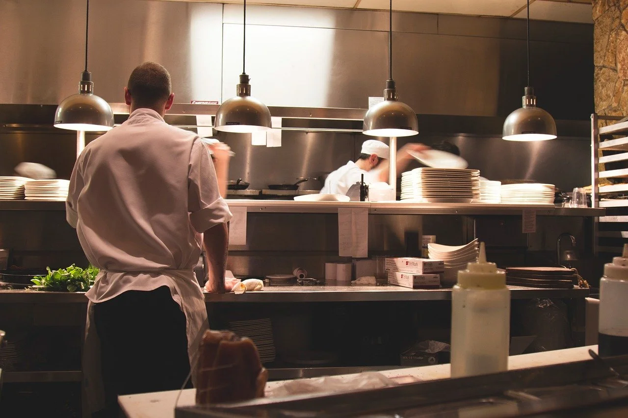 View of a busy restaurant kitchen with chefs preparing food, shelves of plates, and cooking utensils under hanging lights.
