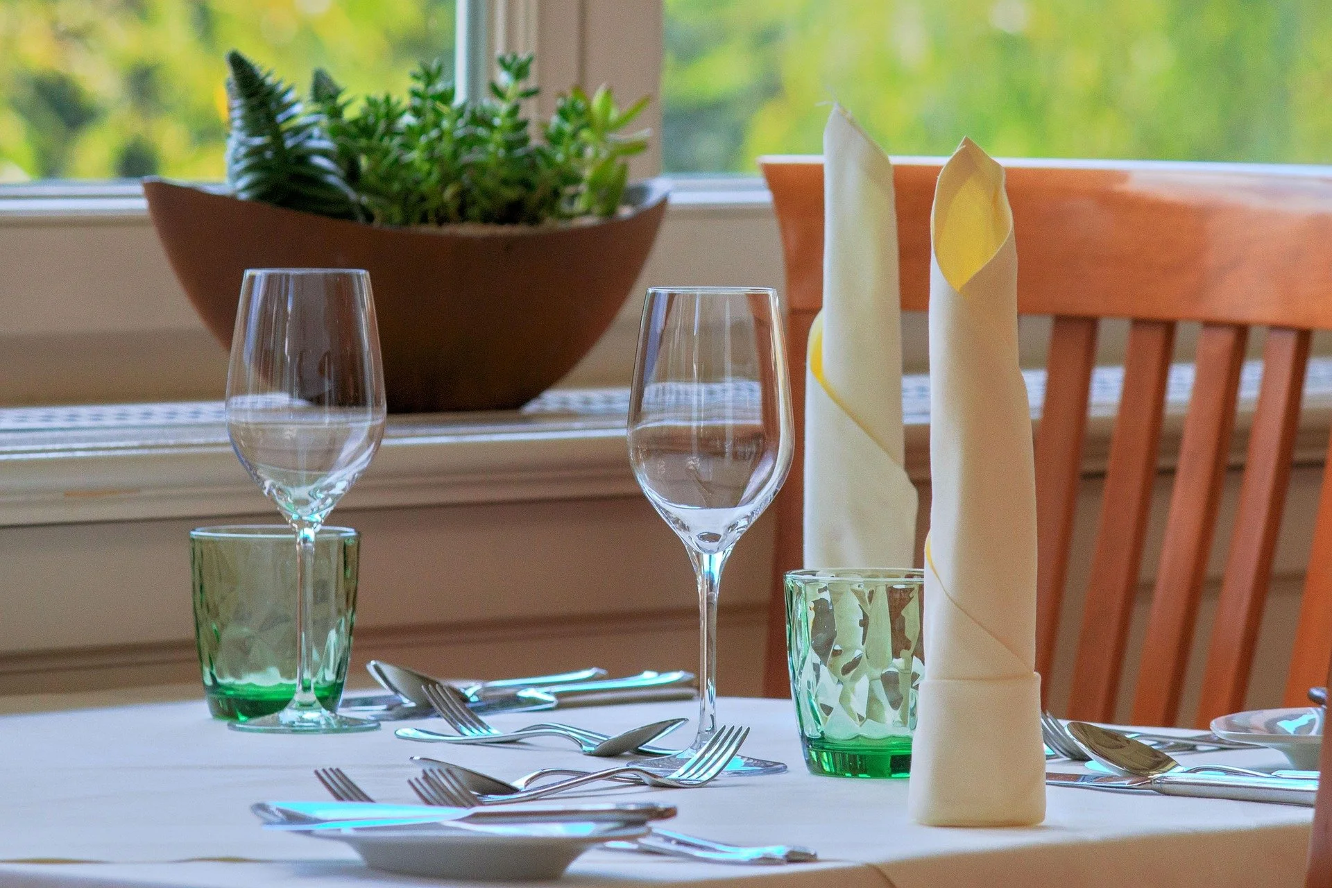 A set dining table with two empty wine glasses, a green glass, a folded napkin, and cutlery on a white tablecloth. In the background, there is a window with a view of greenery outside and a wooden chair.