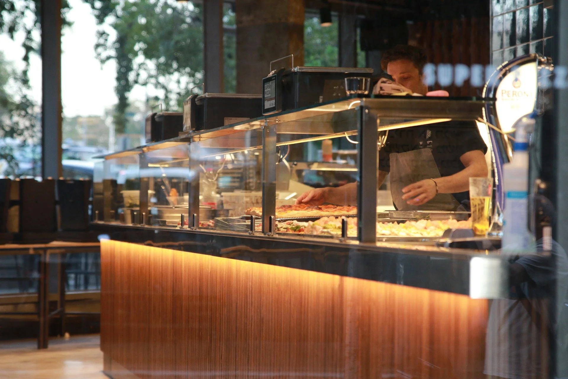 A person preparing food behind a glass display case in a restaurant, with various dishes and a glass of beer on the counter.