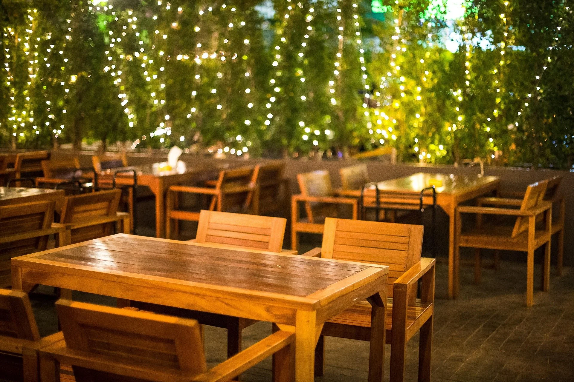 Empty outdoor dining area with wooden tables and chairs, decorated with string lights and greenery in the background.