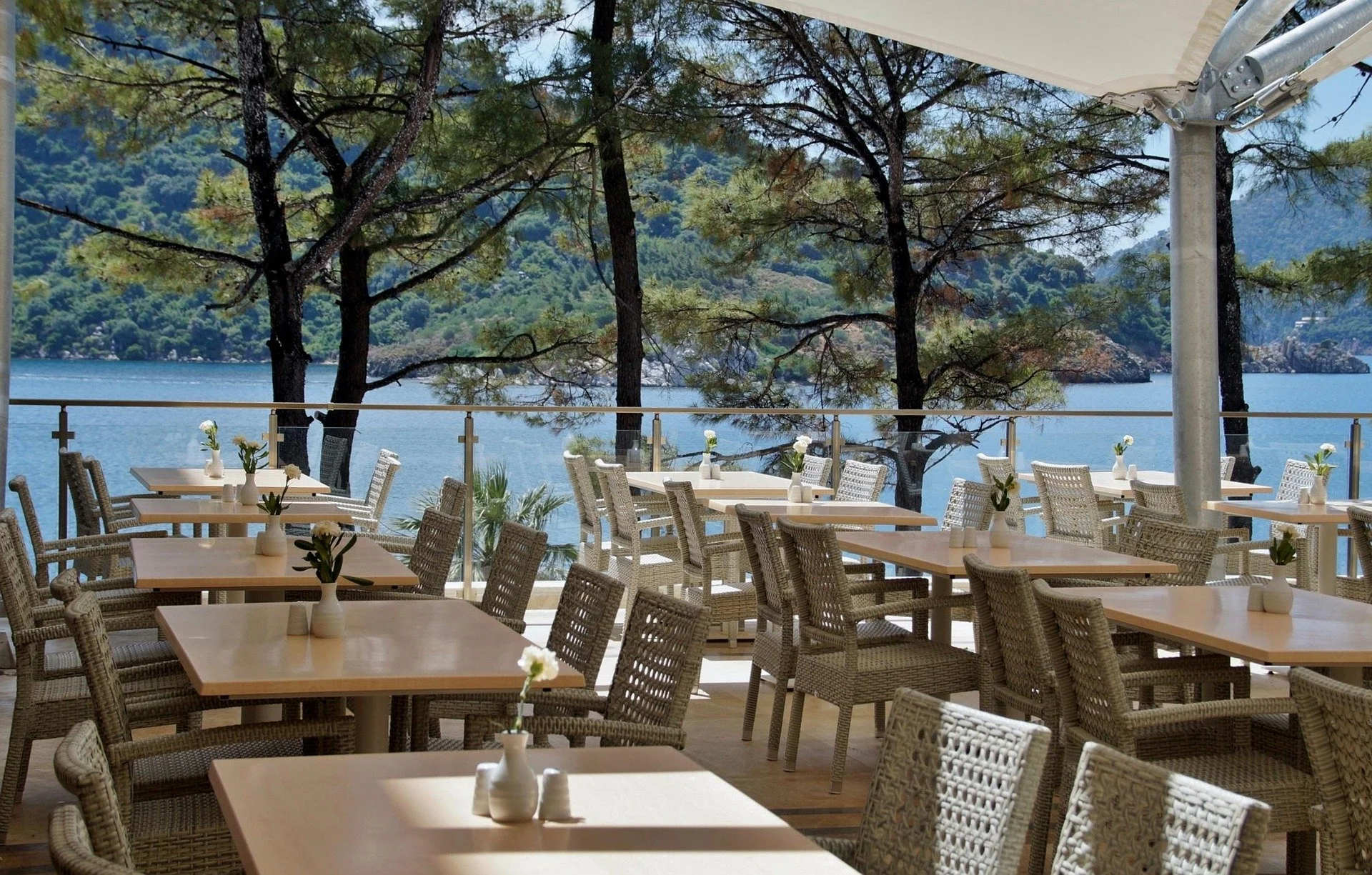 Outdoor restaurant with woven chairs and wooden tables, each decorated with small white vases holding white flowers, overlooking a lake with trees and hills in the background.