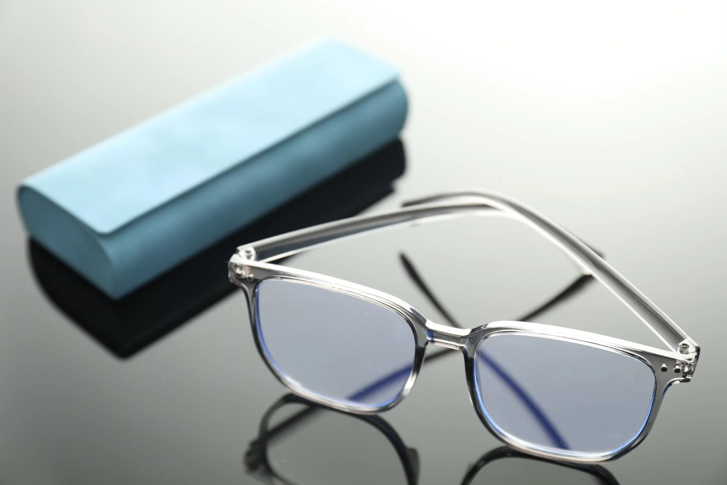 Pair of sunglasses with clear frames and tinted lenses, placed on a reflective black surface, with a light blue and black glasses case in the background.