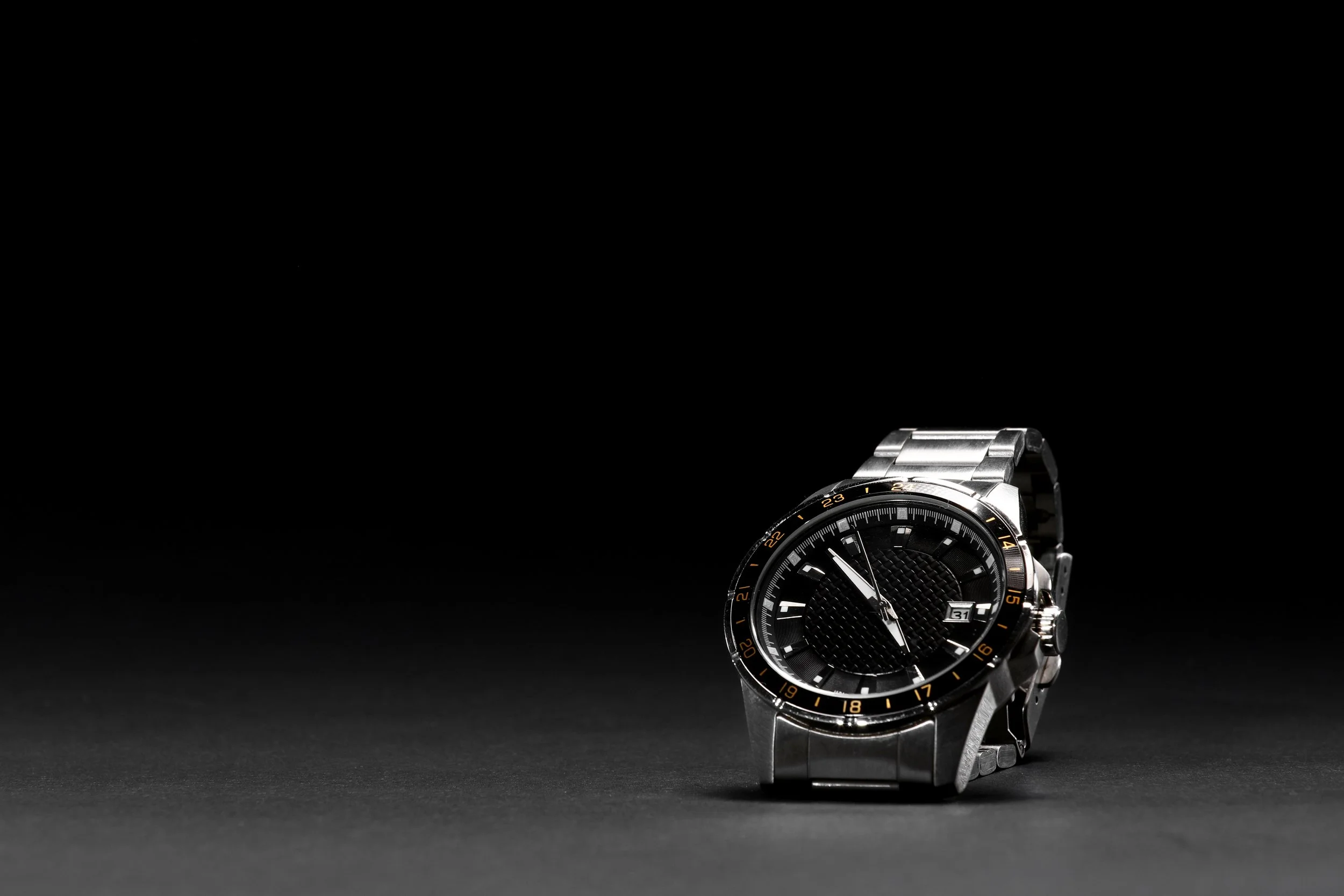 A stainless steel wristwatch with a black face, gold accents, and a date display, placed on a dark surface with a stark black background.