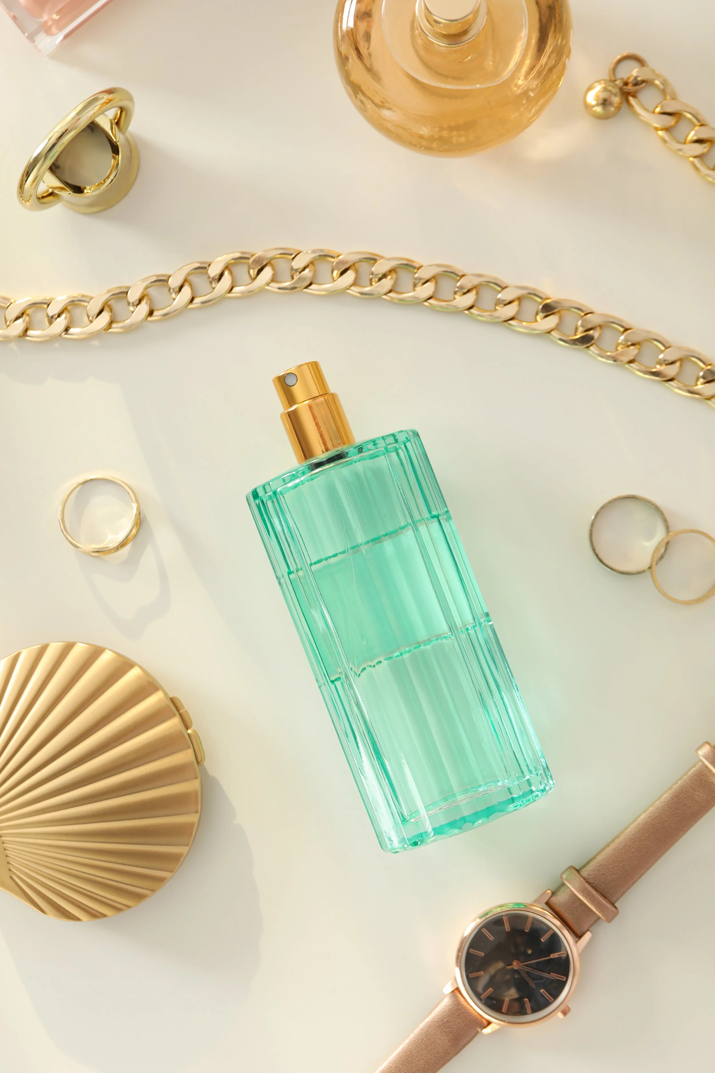 A flat lay of gold jewelry, a green perfume bottle, a gold watch with a brown leather strap, and a glass perfume container on a light surface.