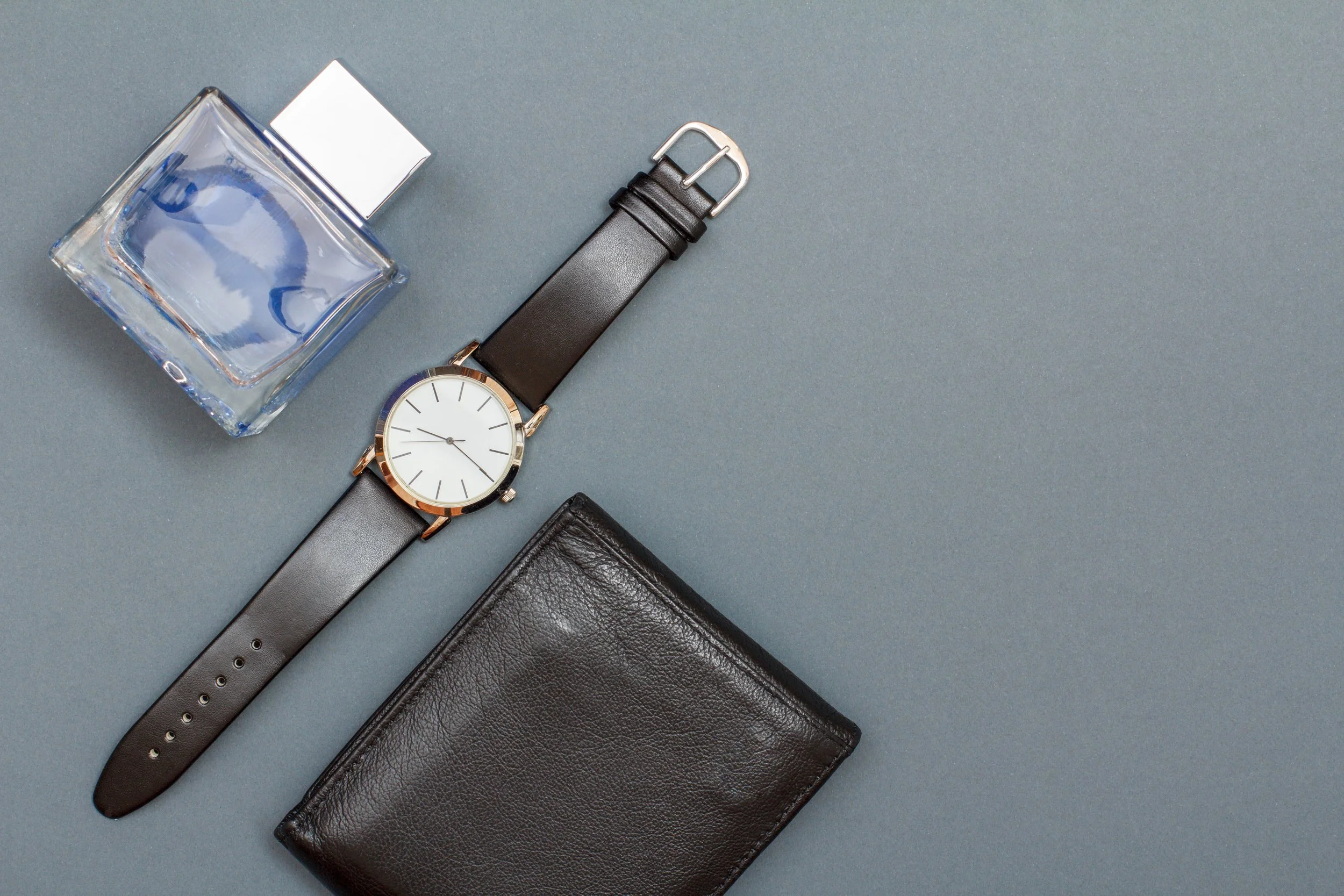 A flat lay of a glass perfume bottle, a wristwatch with a black leather strap and a white face, a black leather wallet, on a gray surface.