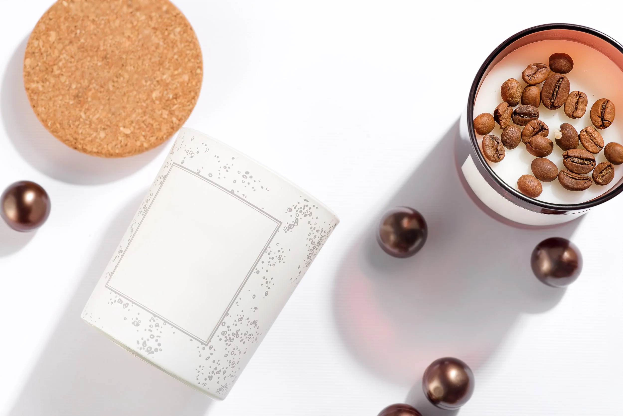 A candle with coffee beans on top, a white candle container with a blank label, a cork coaster, and several bronze-colored decorative balls on a white surface.