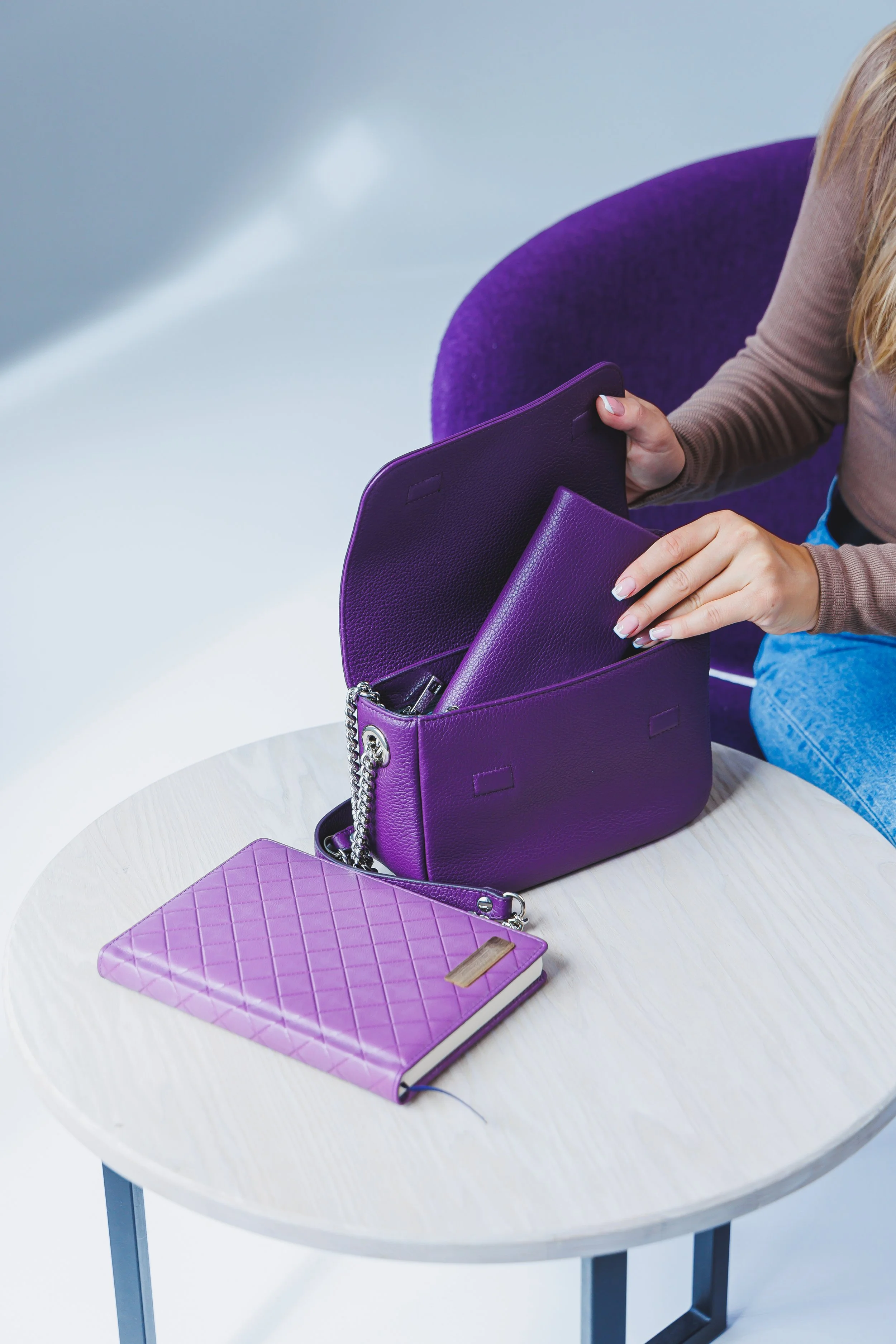 A woman placing a purple wallet into a purple handbag on a light-colored round table. The table also holds a purple quilted notebook with a gold nameplate.