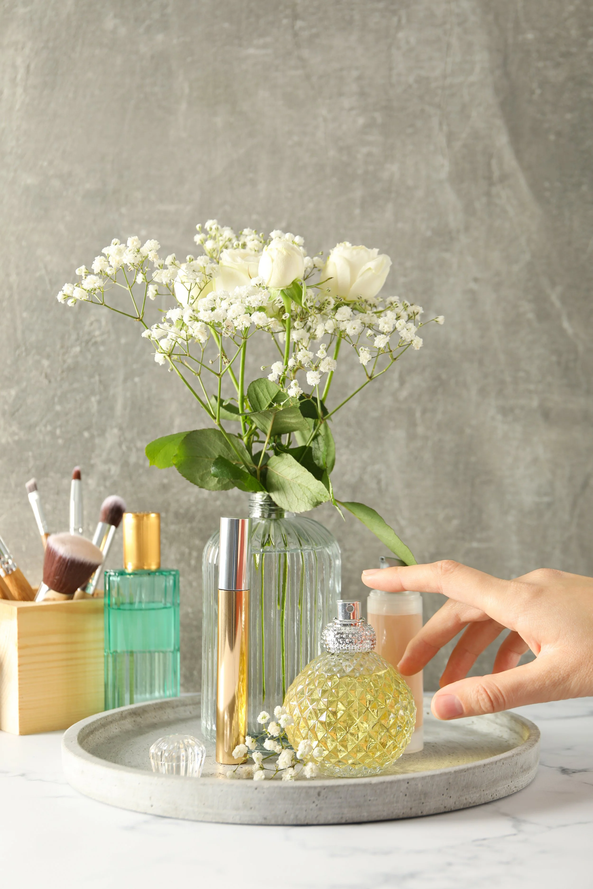 A tray with various perfume bottles, a hand reaching for a yellow perfume, a glass vase with white roses and baby's breath, and makeup brushes in a wooden container on a white marble surface.