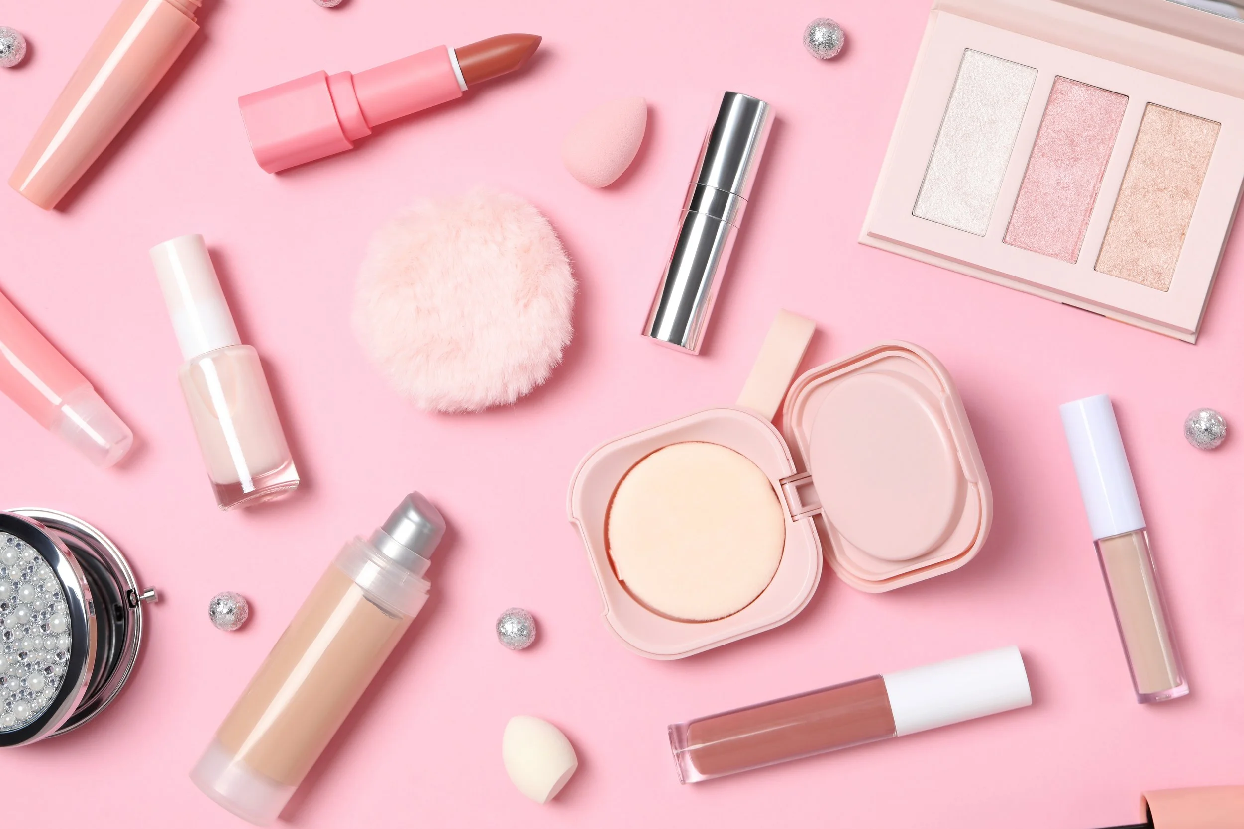 Various makeup products and tools on a pink background, including foundation bottles, a powder compact, eyeshadow palette, lipsticks, brushes, and decorative silver balls.